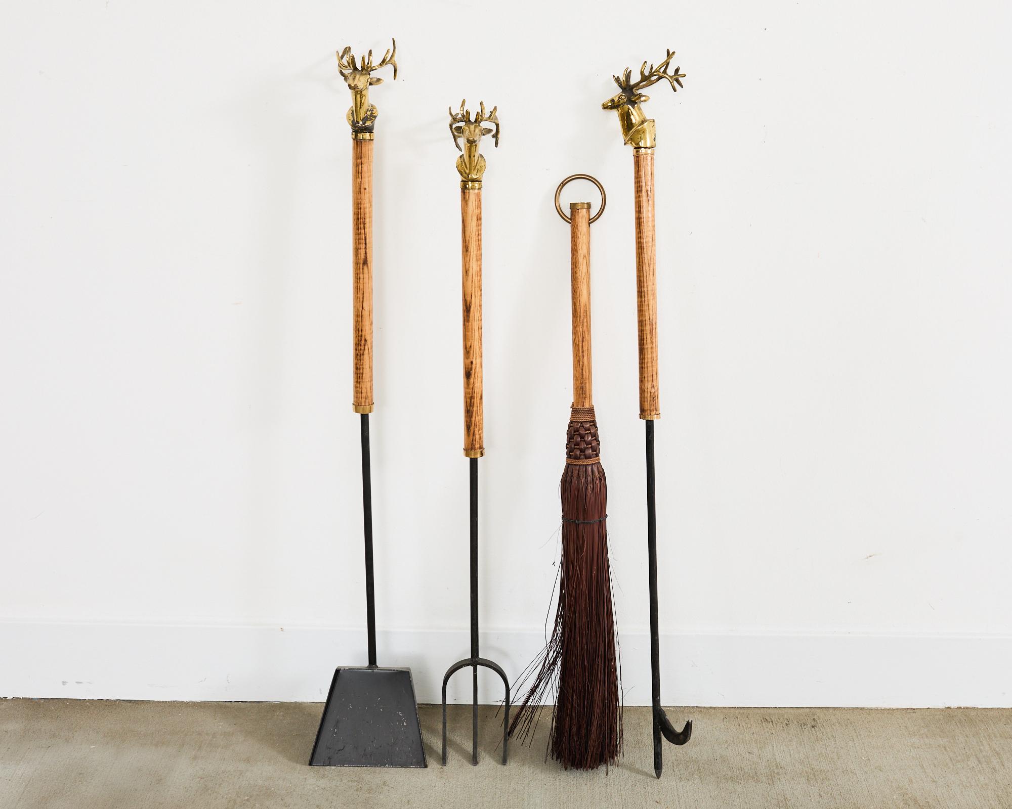 Rustic set of hand-crafted iron fireplace tools and holder featuring patinated brass deer head busts. The set includes a poker, a shovel, a fork, a broom, and a matching holder. The tools have an iron construction with oak handles. The broom or