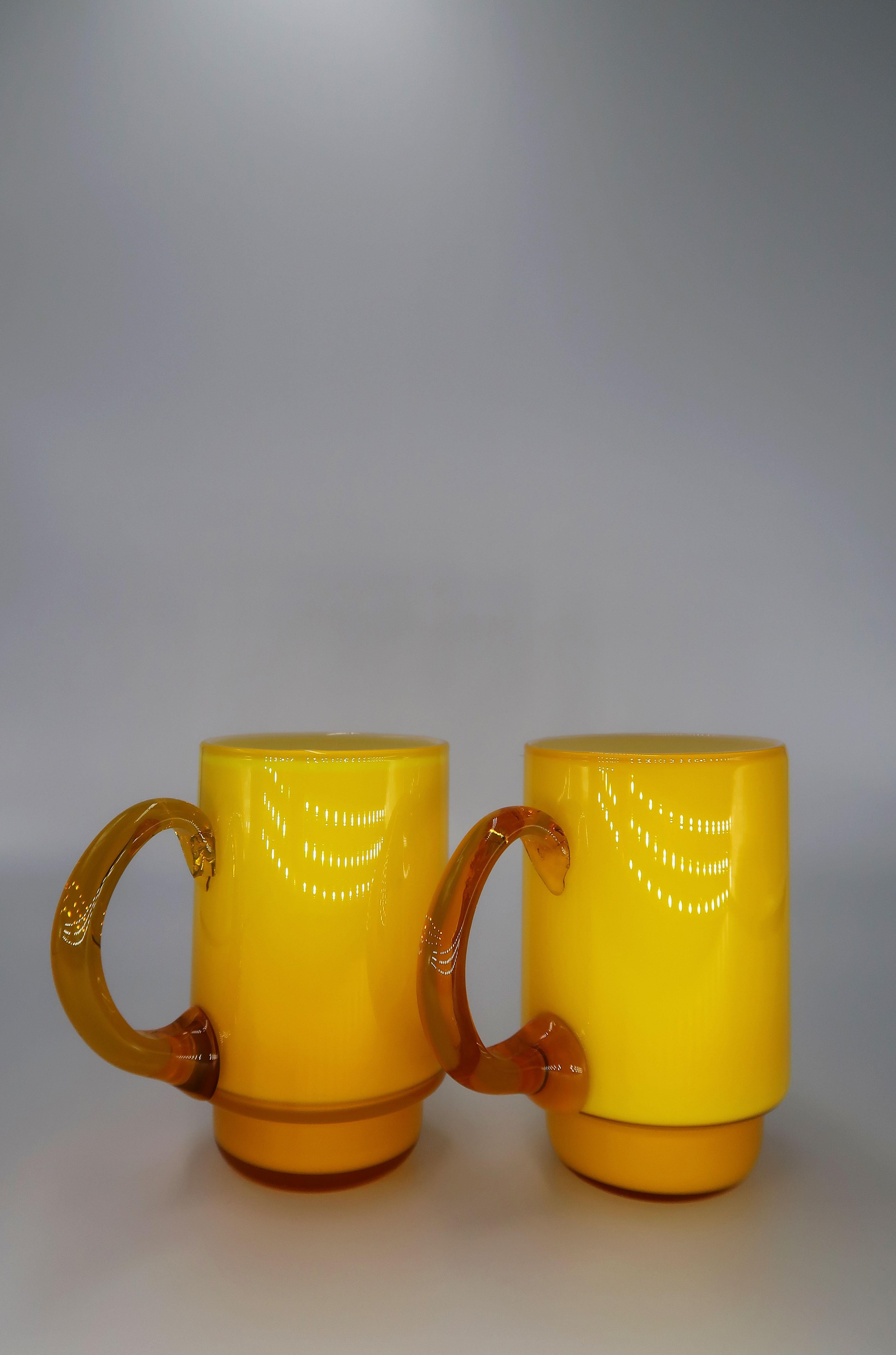 Set of two Danish Mid-Century Modern mouth blown glass cups by acclaimed glass designer Michael Bang (1942-2013). Bright yellow glass on the outside, pure white glass on the inside and caramel handle attached on one side. Due to the handmade nature