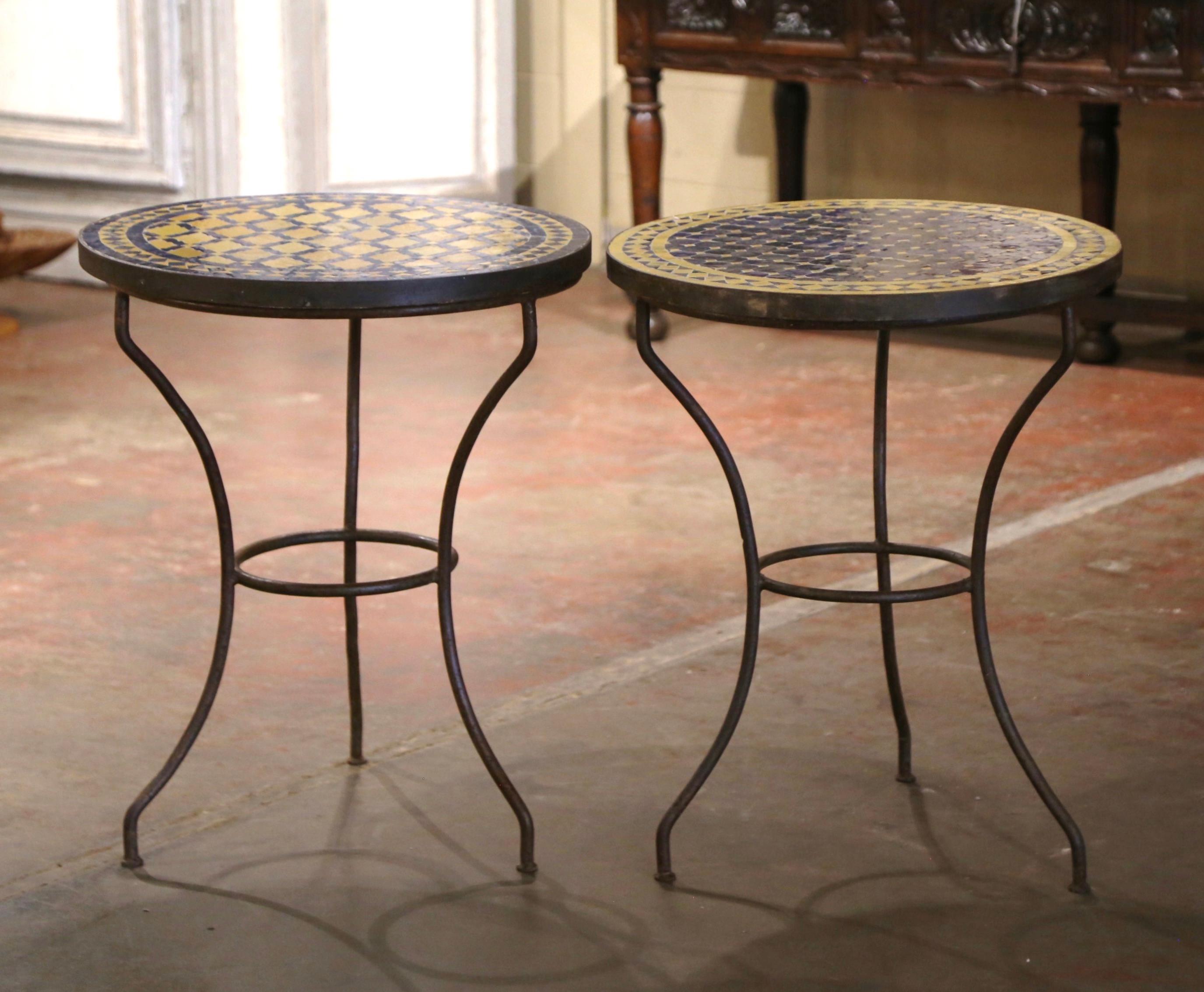 Decorate a patio or covered porch with this colorful set of two of antique bistrot tables with mosaic tops. Crafted in France circa 1920, each table sits on three iron scrolled legs connected with a round center stretcher. One tabletop features a