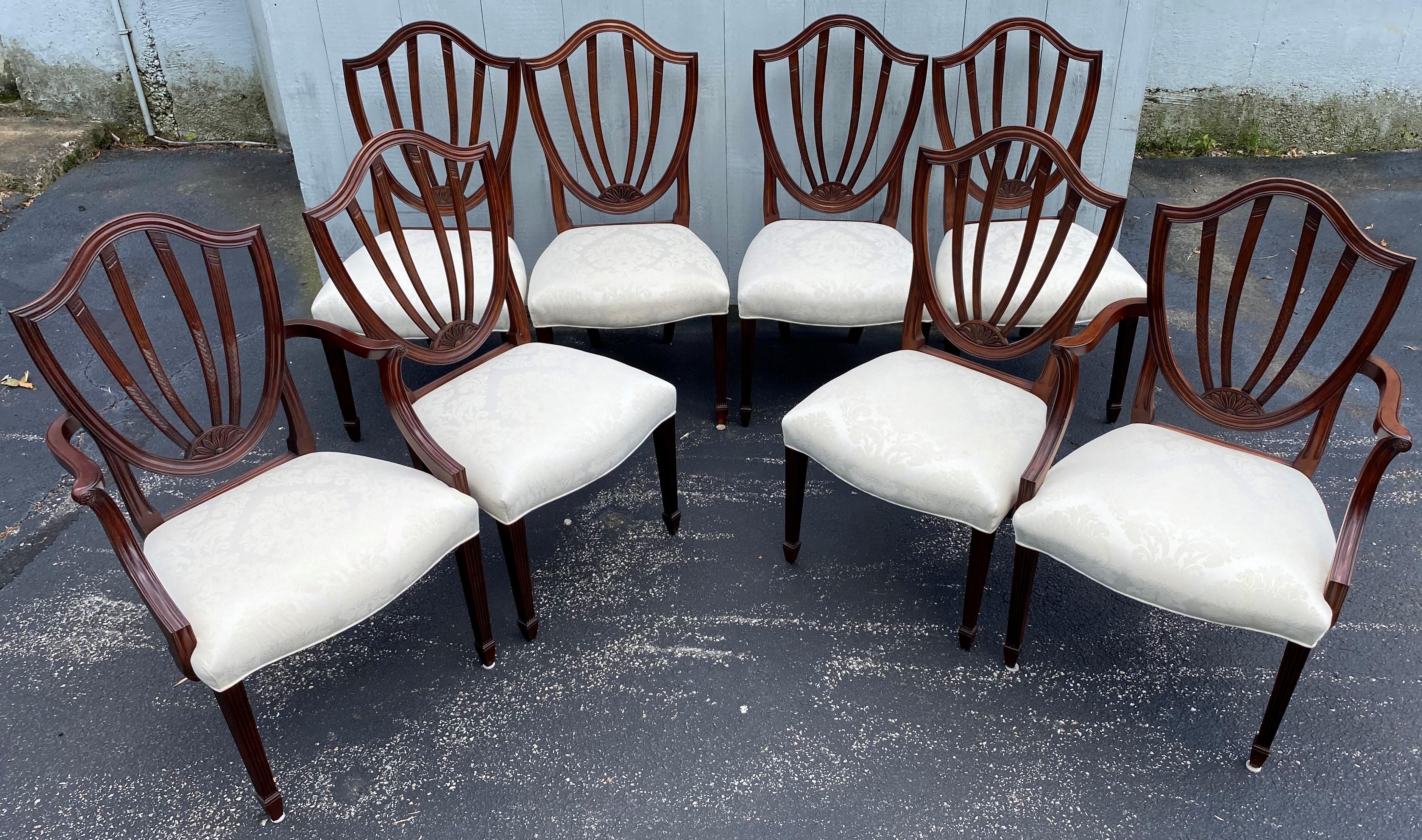 A fine set of six mahogany shield back dining chairs with white damask upholstered seats by Baker Furniture, with detailed foliate carving on the shield back bases, as well as carved and reeded front legs terminating with spade feet. A Baker