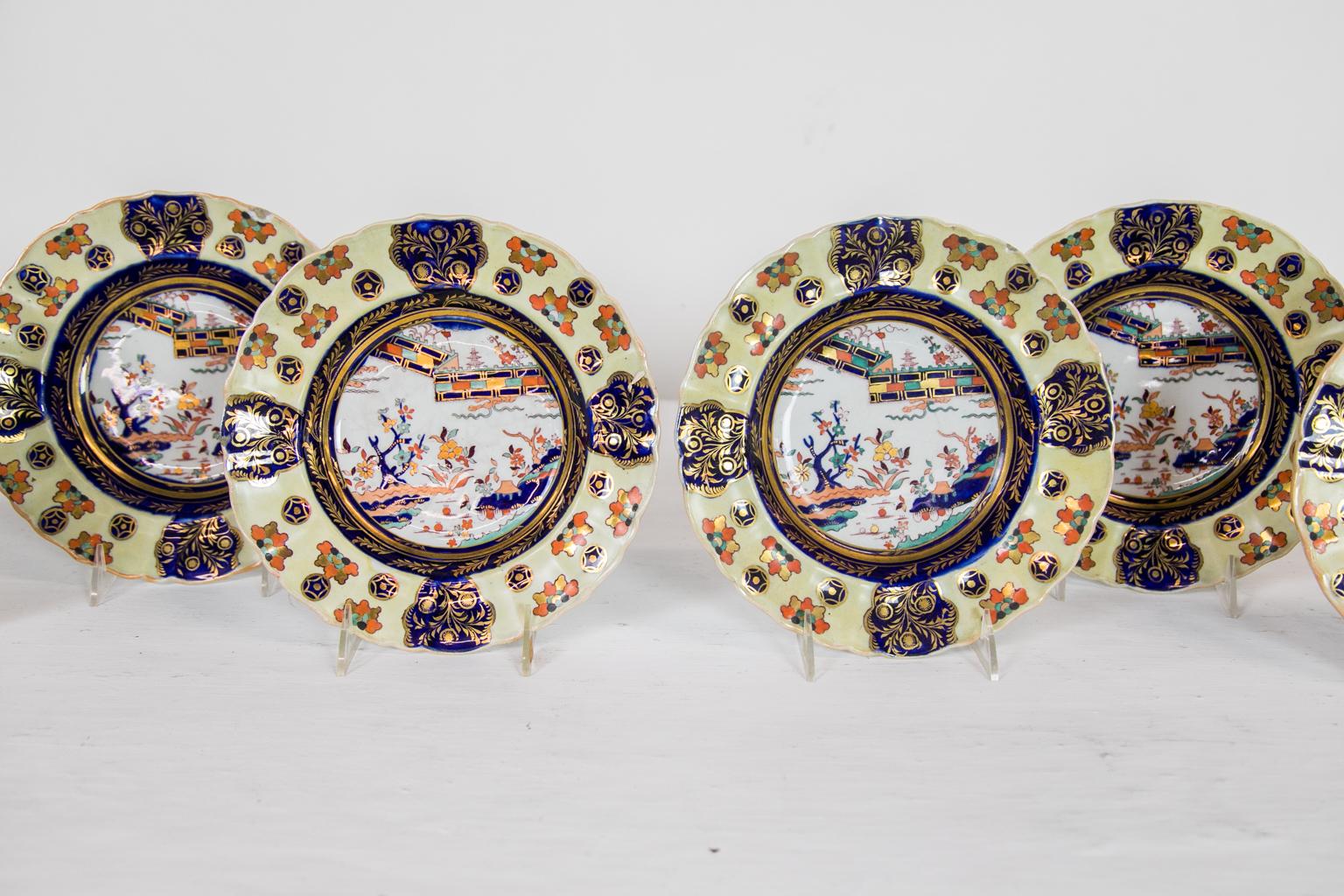 These soup plates have scalloped edgeas that frame a border with a rare yellow background containing stylized leaf clusters and round star lozenges interspersed with four cobalt cartouches of gilt leaves. The center panel has a fence pattern and a