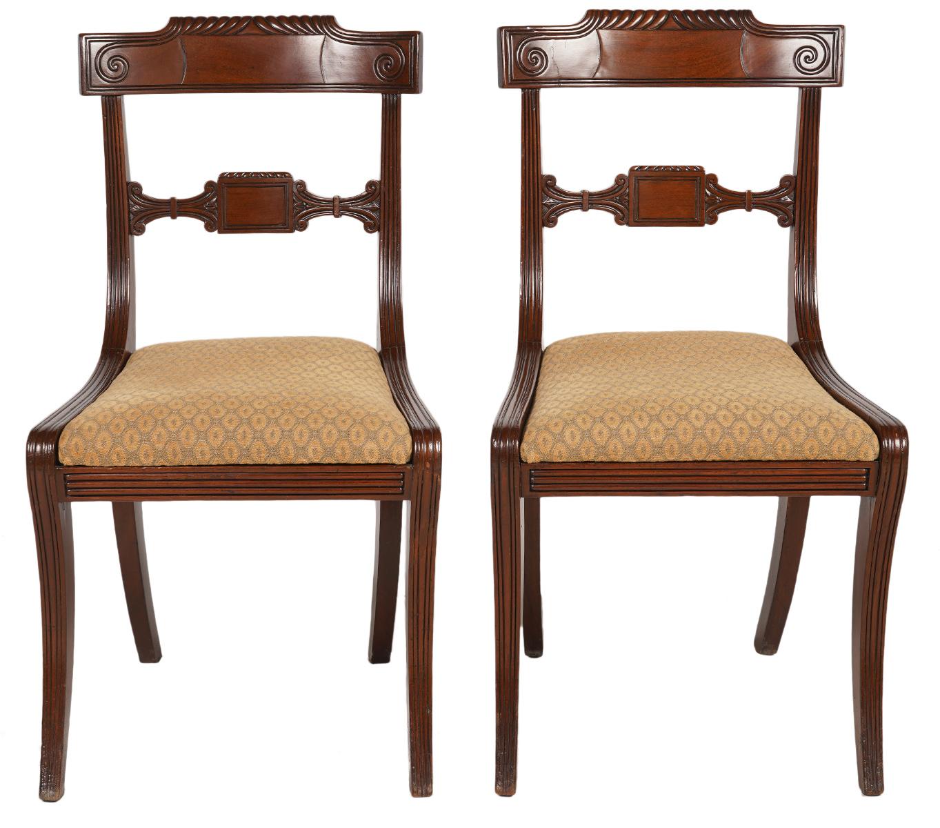 These English Regency dining chairs in the Klismos style feature carved, gadrooned and curved back rails and splats. The frames are beautifully reeded and the armrests on the armchairs ends in large scrolls. The seats are upholstered and covered