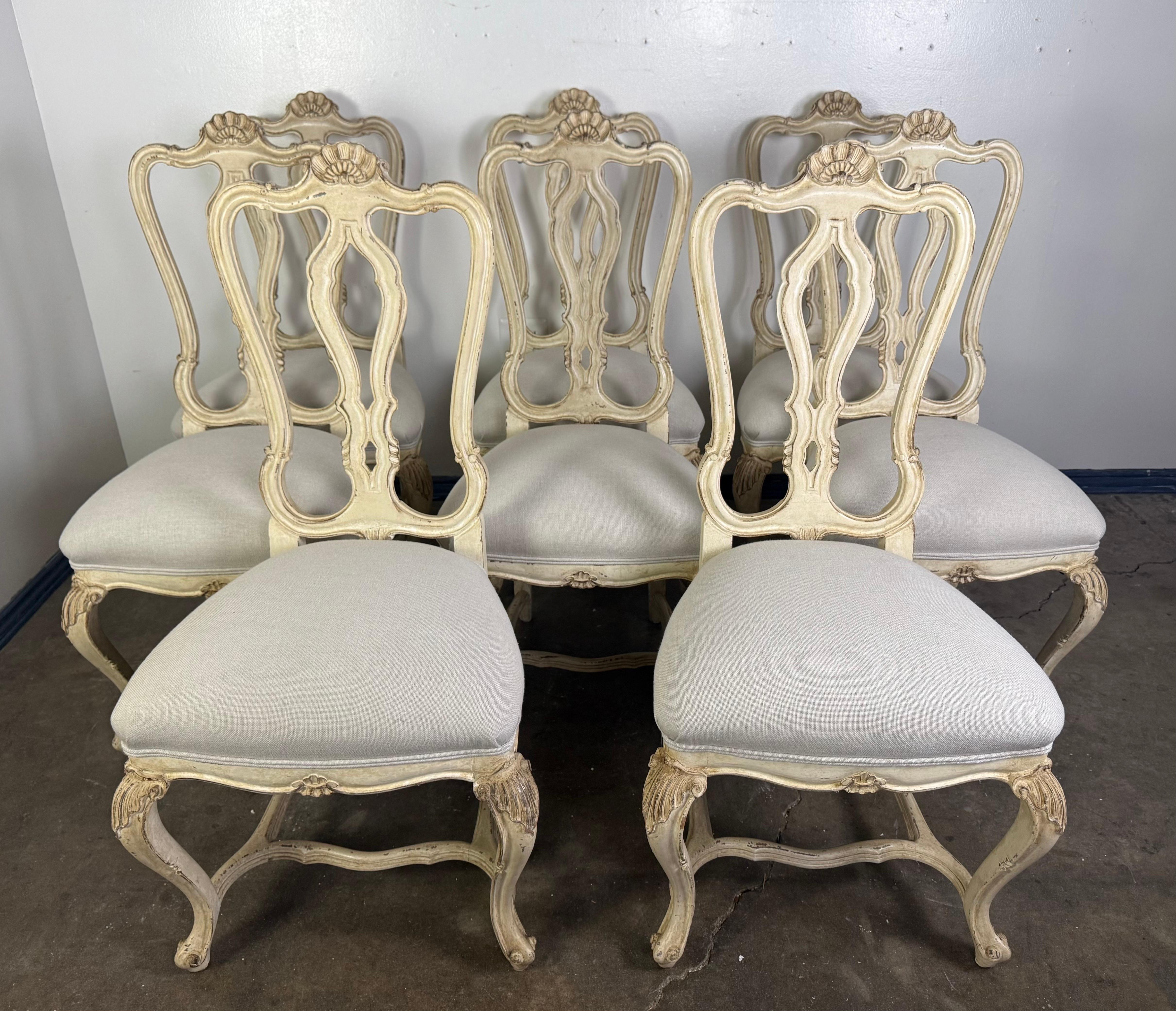 
This elegant set of eight dining chairs captures the graceful lines and refined ornamentation of the Louis XV style. Each chair features a hand-carved wooden frame with a beautifully aged, painted finish in a soft, creamy tone with subtle