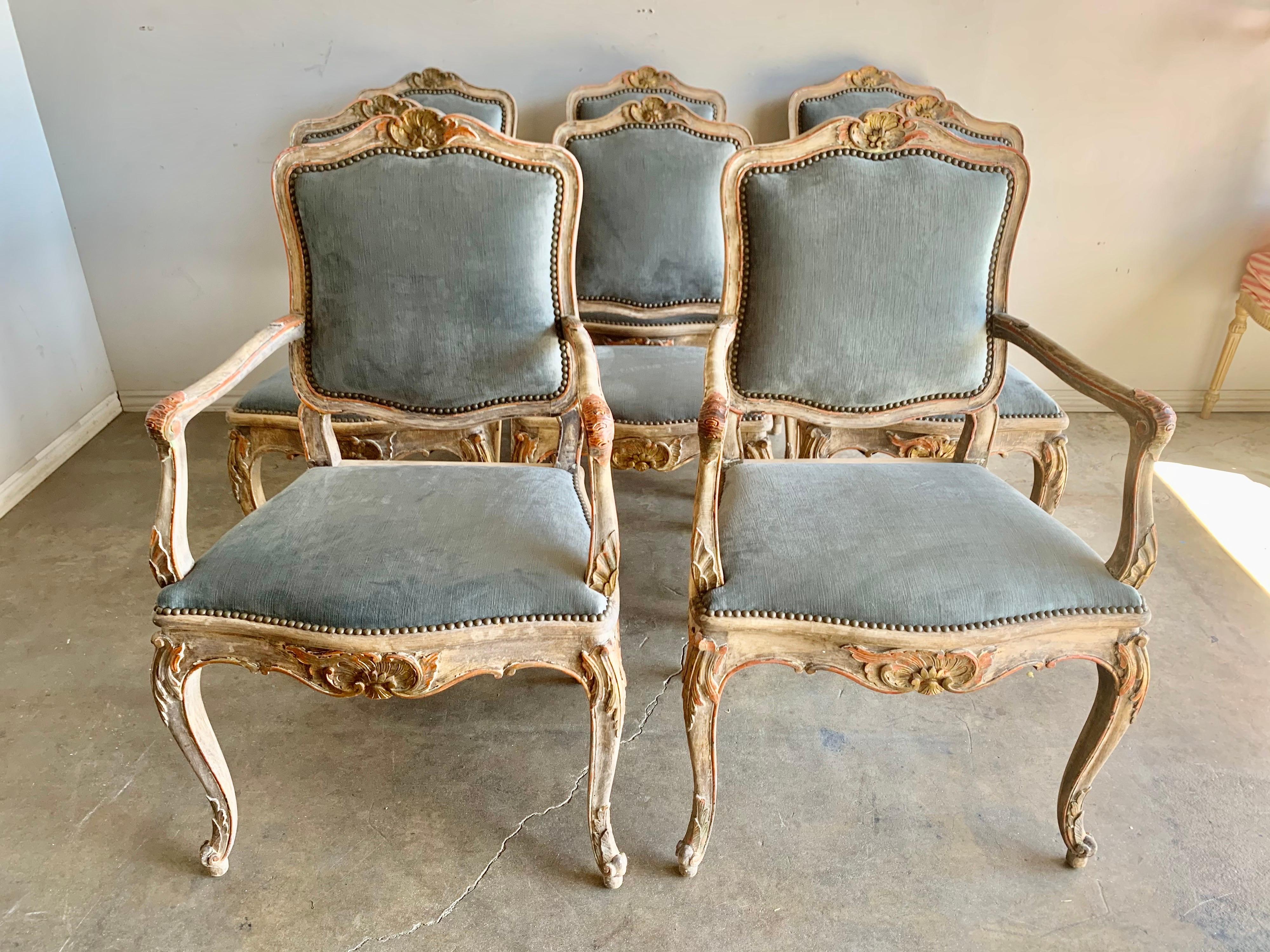 Set of eight carved French Lois XV style dining chairs with a painted and parcel gilt finish. The chairs stand on four cabriole legs with rams head feet. The chairs are newly upholstered in a steel blue velvet and detailed with antique brass