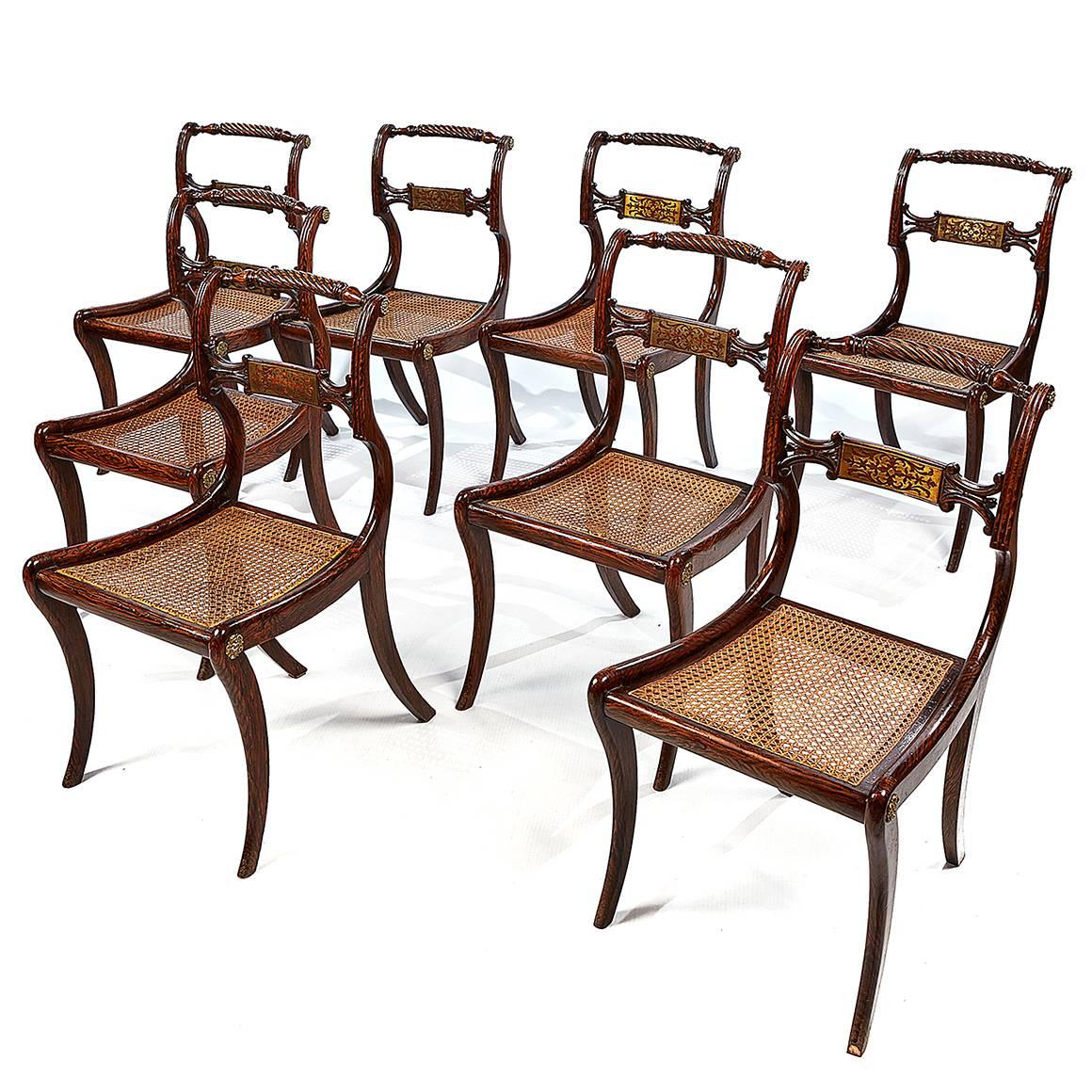 Each dining chair in this set of eight features a simulated rosewood turned and carved rope-twist crest rail ending in brass rosettes, above a brass-inlaid back rail. With a cushion atop a cane seat and sabre legs.