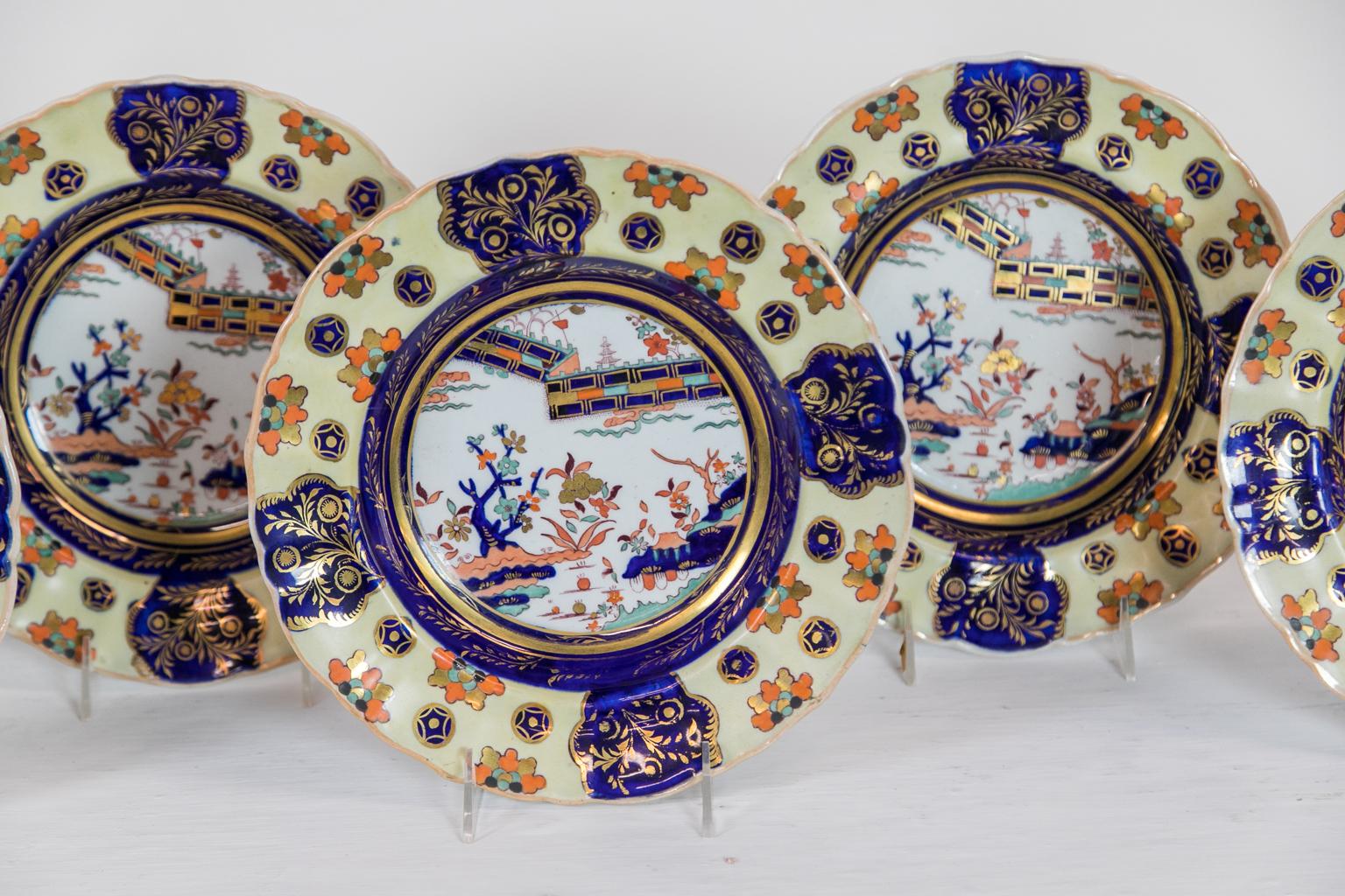 These soup plates have scalloped edges that frame a border with a rare yellow background containing stylized leaf clusters and round star lozenges interspersed with four cobalt cartouches of gilt leaves. The center panel has a fence pattern and a