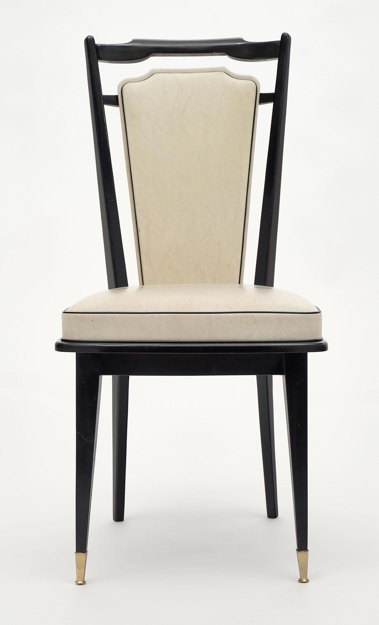 A set of eight chairs made of solid mahogany with legs capped with brass feet. The wood has been ebonized and finished with a lustrous French polish. They feature their original ivory piped vinyl upholstery.