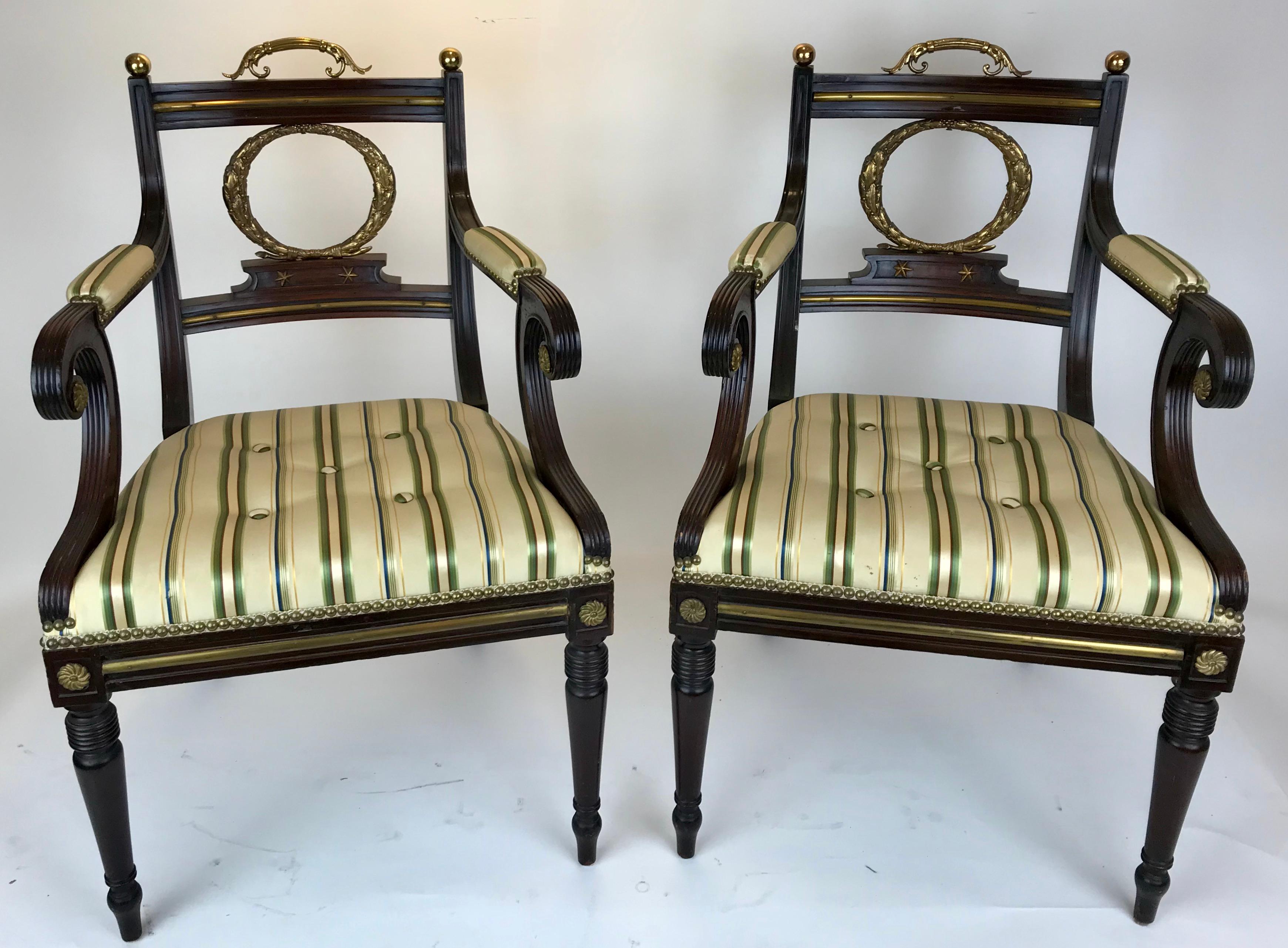 This set of eight English Regency dining chairs include two armchairs with scrolled arms. Each backrest features a gilt bronze lifting handle and oak leaf wreath form splat. The strait front upholstered seats are raised on tapered legs terminating