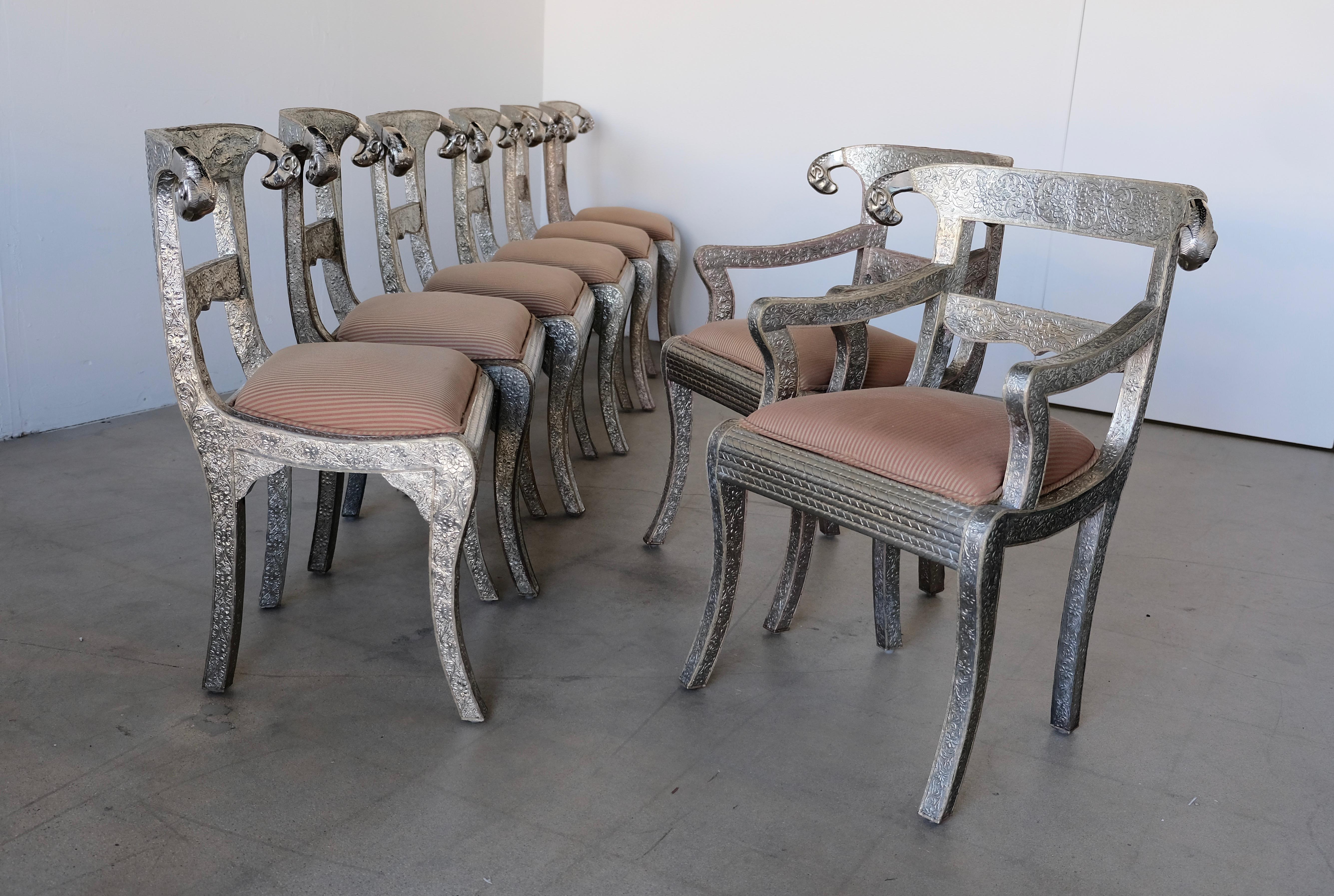 A set of eight Anglo-Indian handmade dining chairs. The chairs are embossed metal over wood finished with curved decorative cast Rams heads. The set consists of six side chairs and two captains chairs with arms. The metal respoussé has a floral and