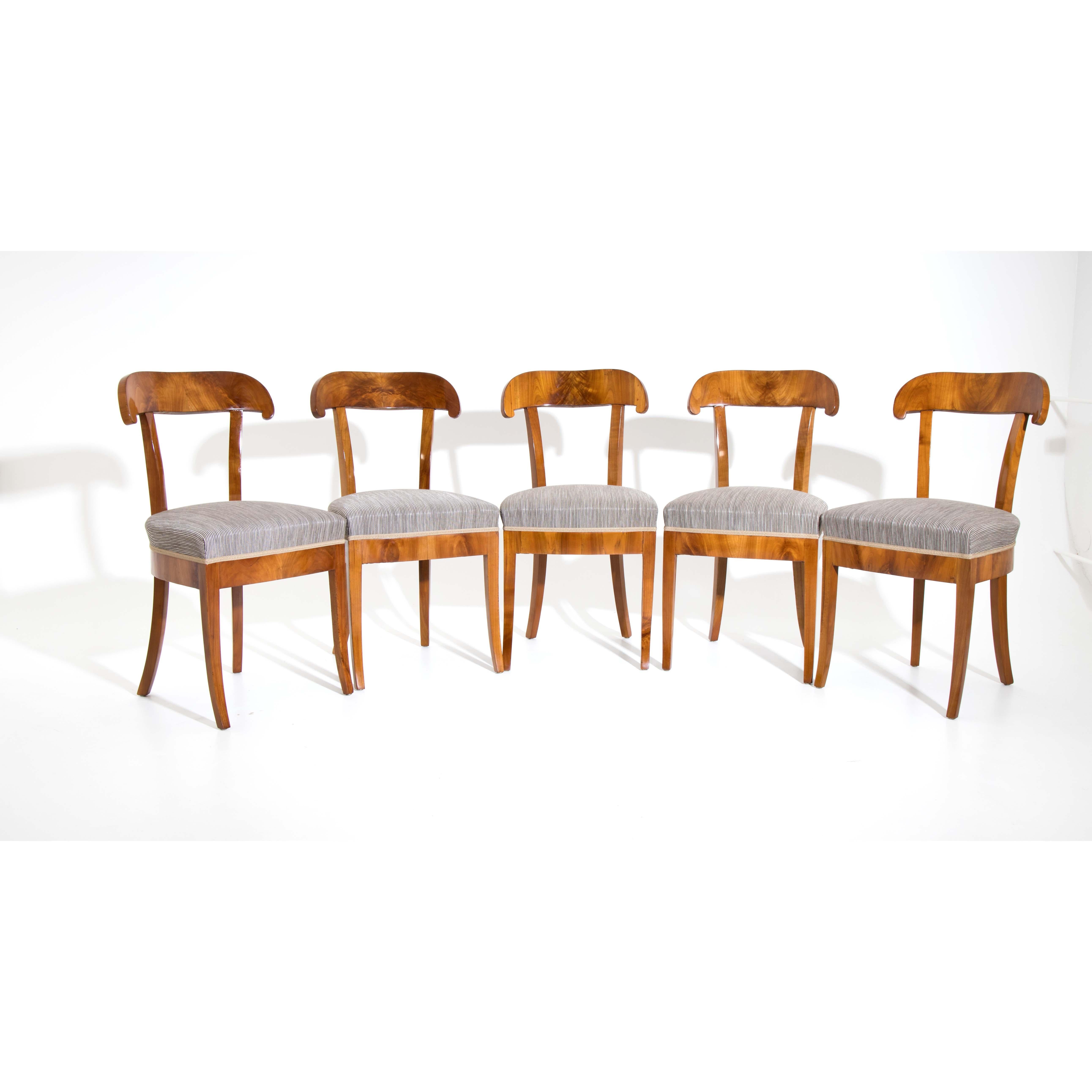 Ensemble de cinq chaises-pelles Biedermeier aux pieds légèrement évasés et aux traverses droites. Les chaises sont plaquées en cerisier. Elles sont nouvellement recouvertes d'un tissu rayé gris et polies à la main.