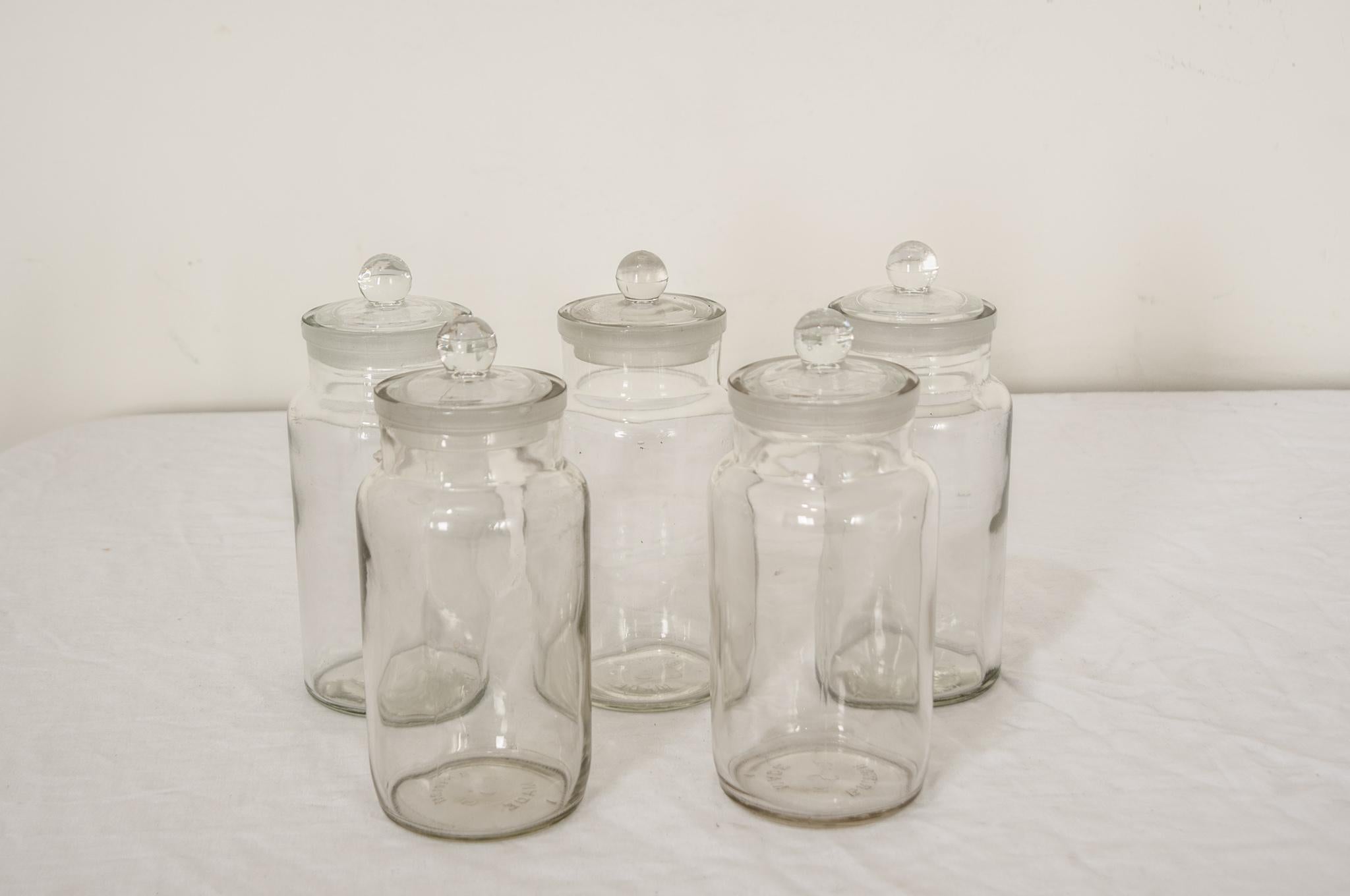 A set of five glass apothecary jars with ball lids, hand-blown in England. Marked by the glassmaker on the bottom with “British S Made”. The antique look of these clear glass apothecary jars make them a beautiful accent in their own right, or an