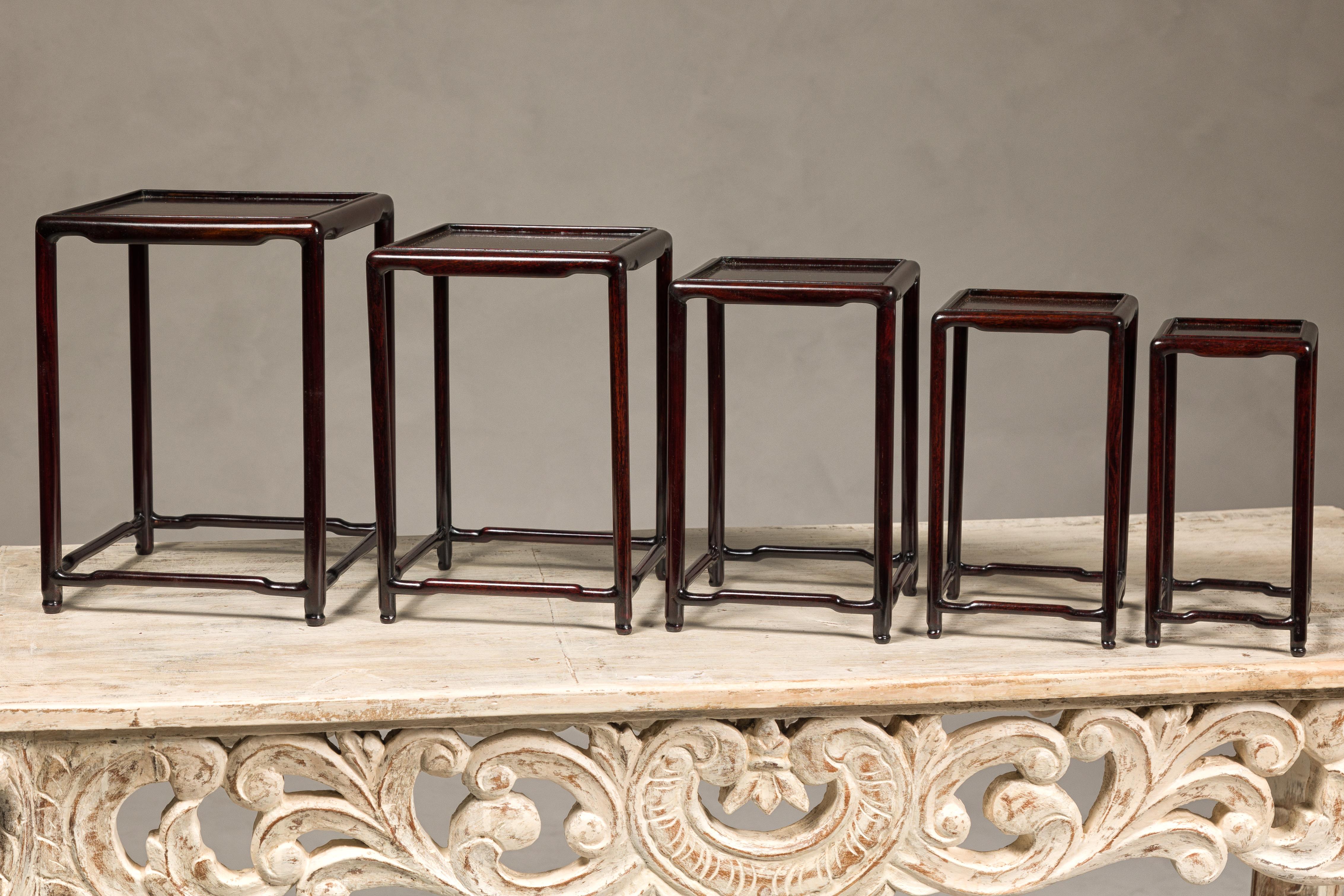 Ensemble de cinq tables gigognes en bois de rose de petite taille, avec des plateaux carrés, des pieds en dos d'âne et une couleur brun rougeâtre foncé. Cet ensemble de cinq tables gigognes en bois de rose du XXe siècle respire l'élégance