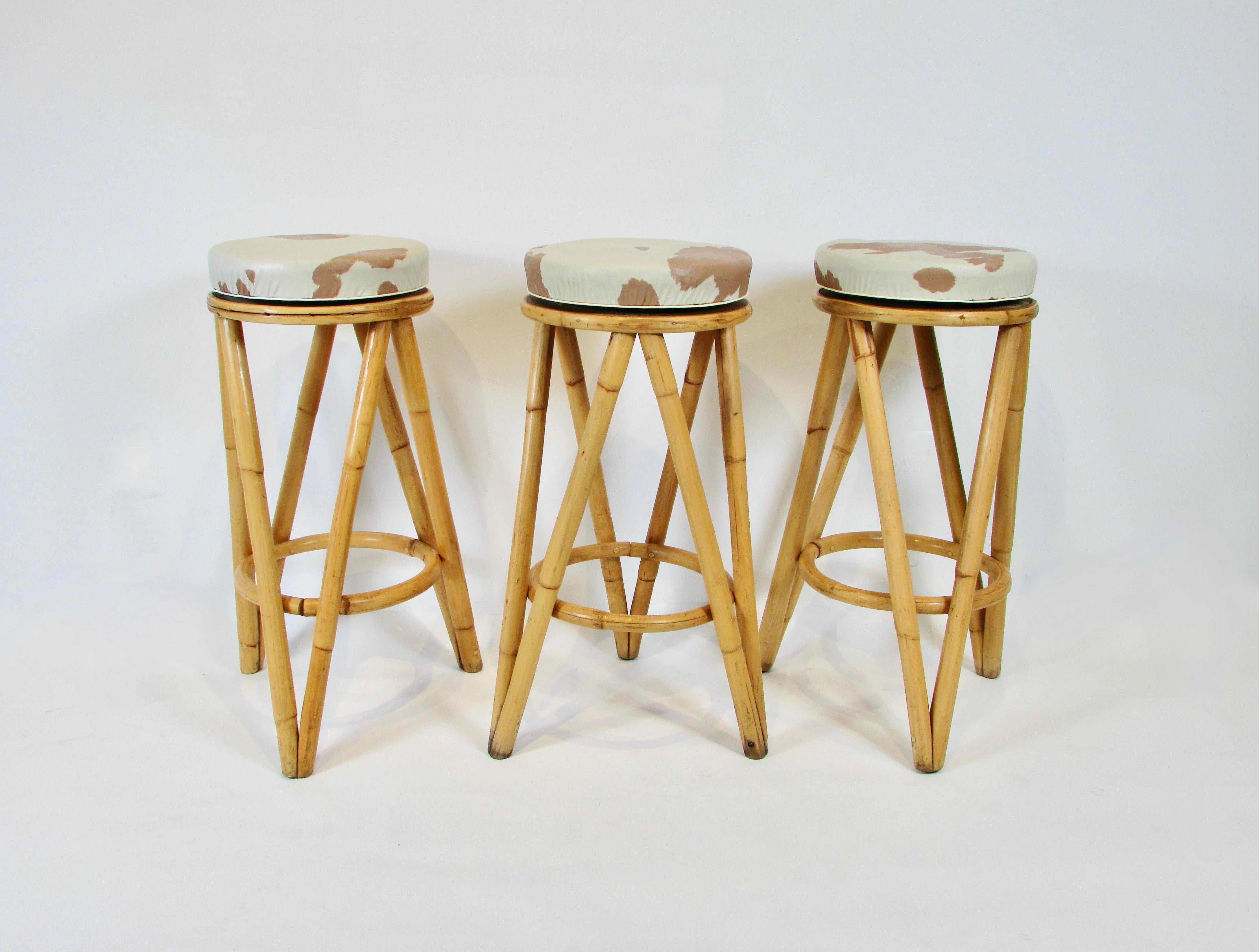 Set of four 1950s bamboo or rattan bar stools . Three triangulated legs support round swivel tops . Seats are covered in what looks like original faux cowhide vinyl . Good condition especially considering age but some wear on seats . Scuffs to