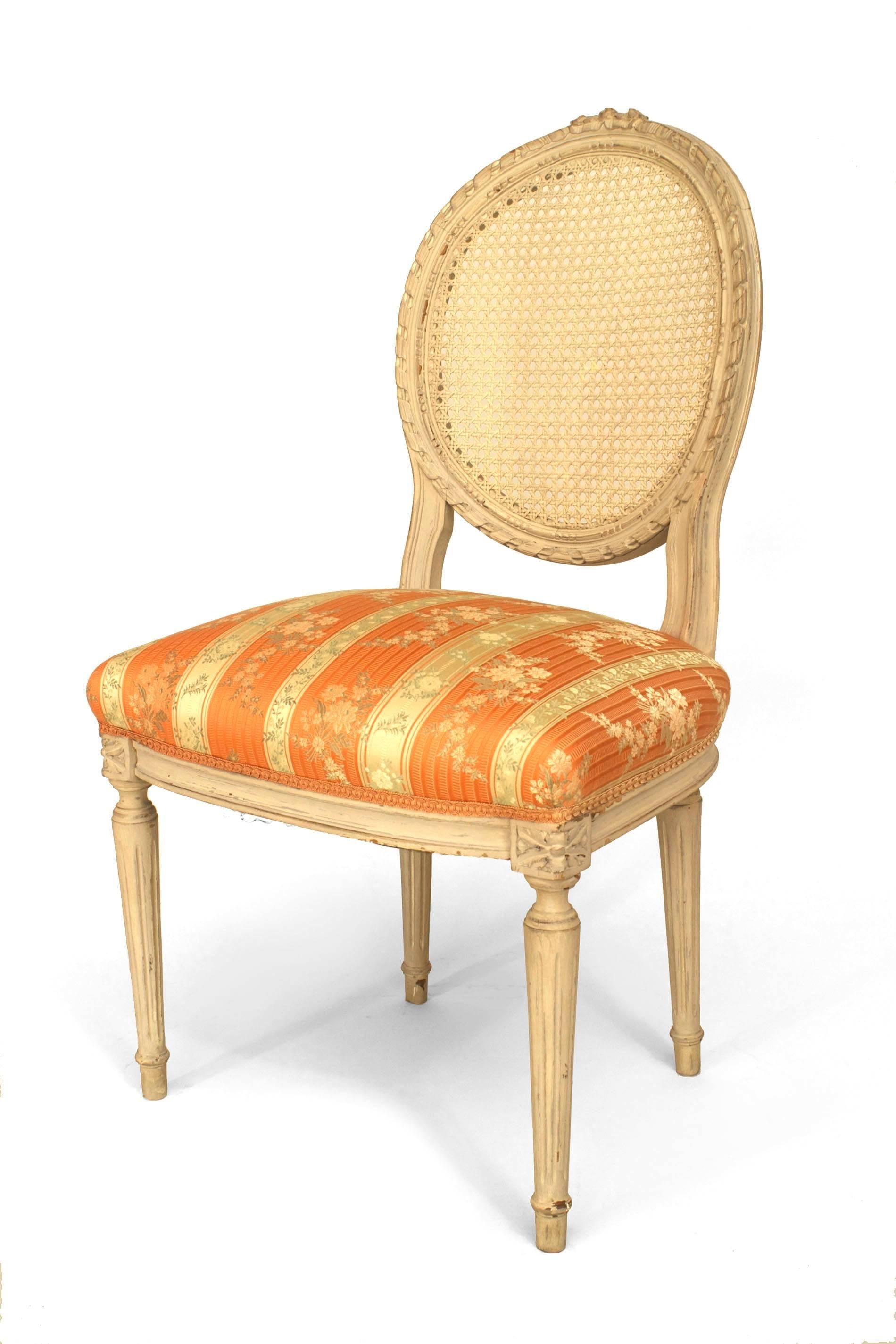 Ensemble de 4 chaises d'appoint de style Louis XVI (20e siècle) peintes à la crème, avec un plateau sculpté en nœud papillon, un dossier ovale en rotin et une assise en damas floral rose et crème.

