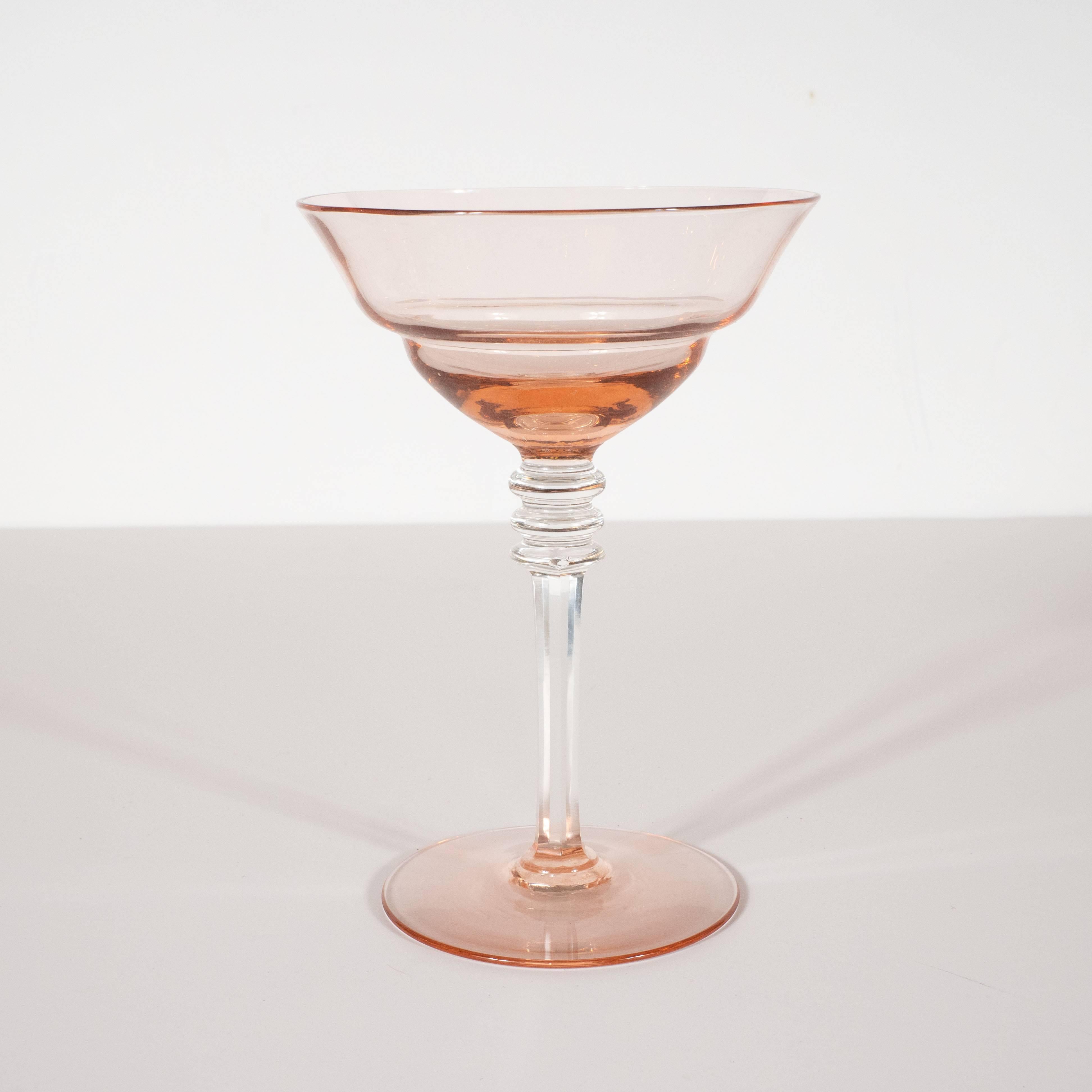 This refined set of four Art Deco drink glasses were realized in the United States, circa 1935. They feature circular bases in a blush rose hue from which a hexagonal stem in clear glass ascends connecting to three circular rings, also in