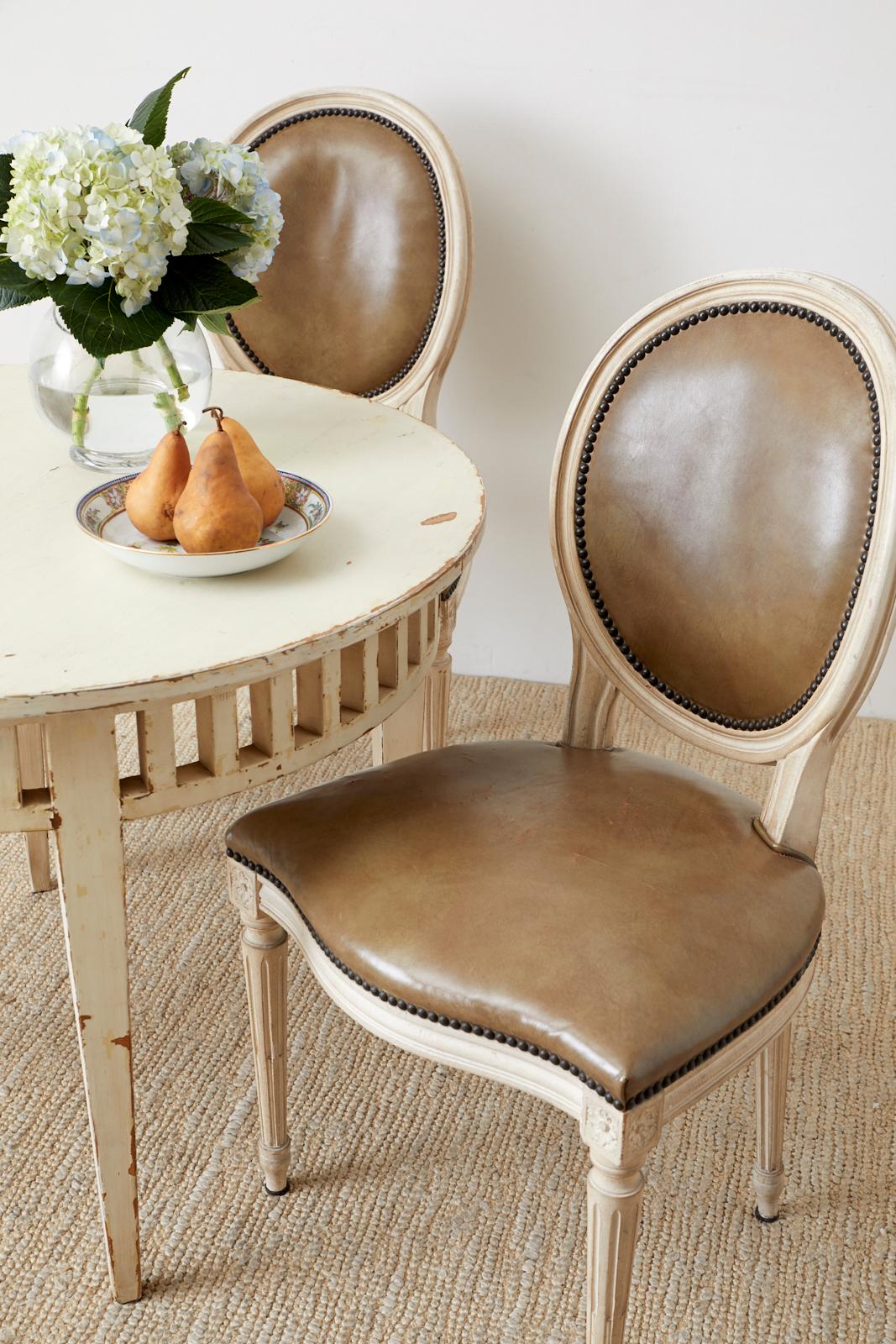 Distinctive set of four Baker furniture dining chairs featuring a painted finish made in the French Louis XVI taste with original leather seats in Baker's satinwood color. The upholstery is bordered with patinated brass tack nail heads to contrast