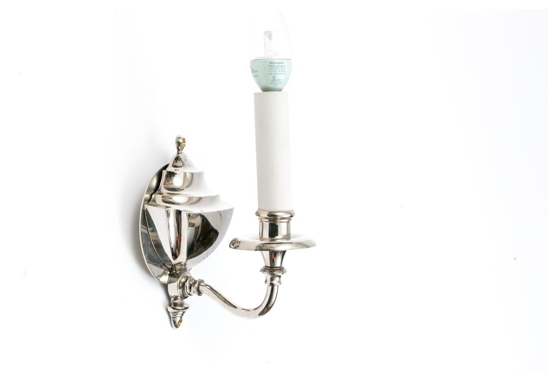 Juego de cuatro apliques de pared electrificados en forma de escudo de cromo pulido sobre latón, con un solo brazo que sostiene una vela de porcelana bizcochada. Los apliques llevan la marca B & H (Bradley & Hubbard). Vienen con placas traseras