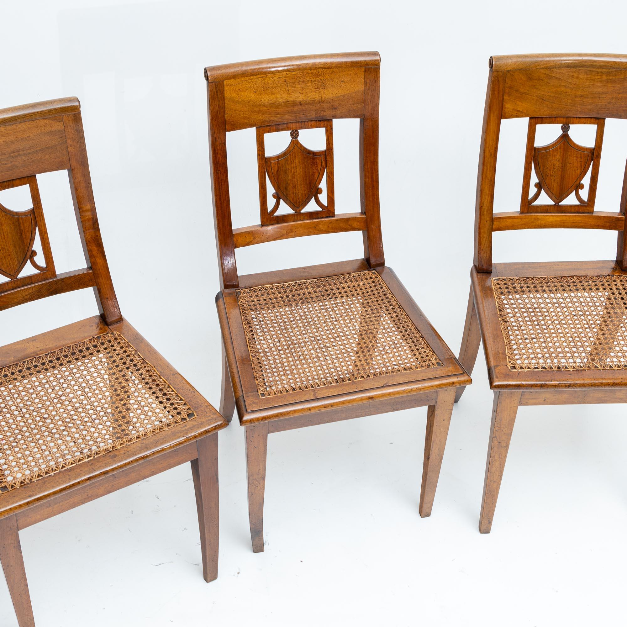 Set of four dining chairs with openwork backs with coat of arms decoration. The chairs stand on square pointed legs with straight frames and a seat covered with wickerwork.