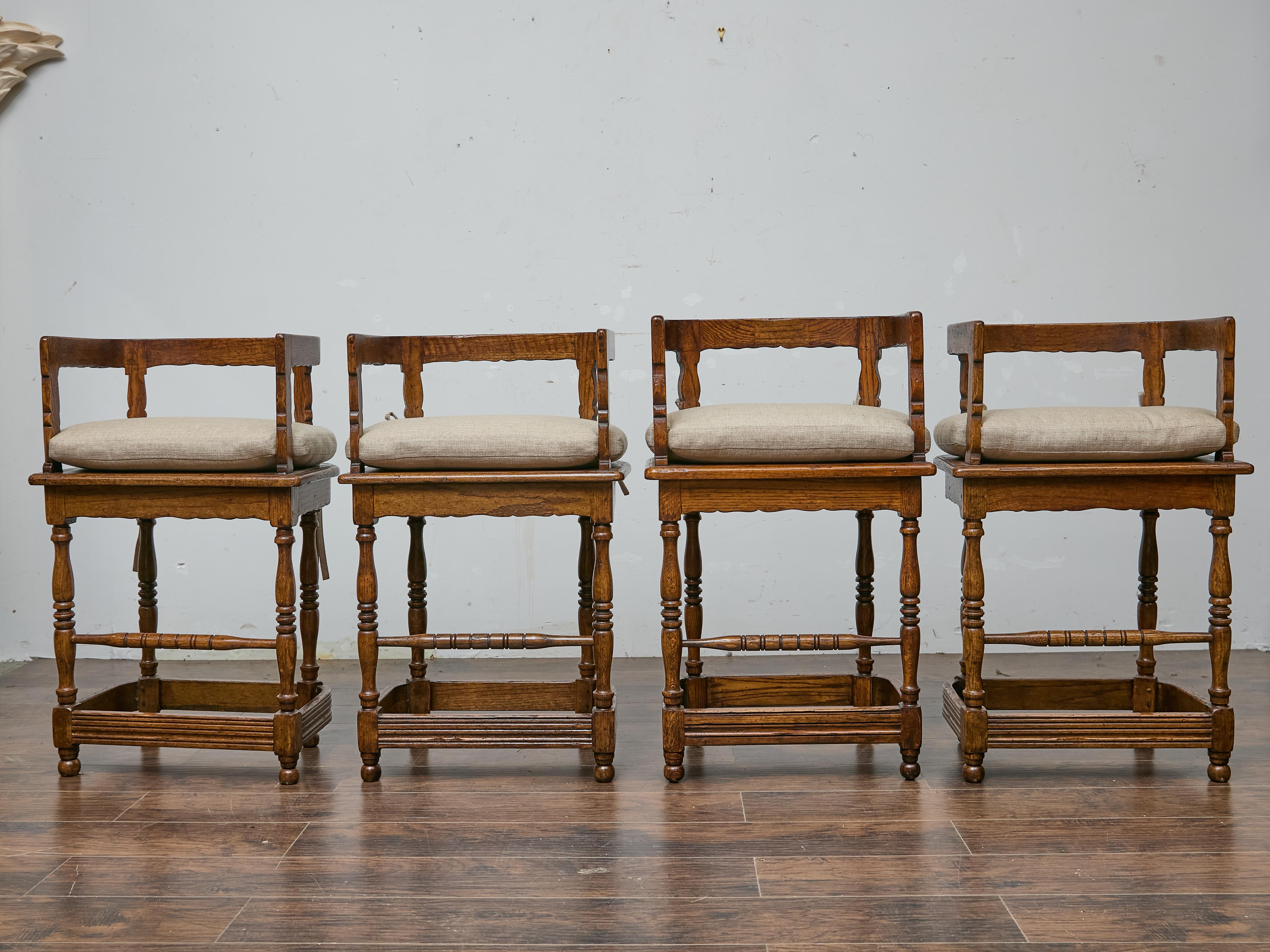 A set of four English oak bar stools from the midcentury period, with rectangular seats and open backs designed for comfort and durability. The frames feature turned legs with ring-turned stretchers doubling as footrests, creating a sturdy and