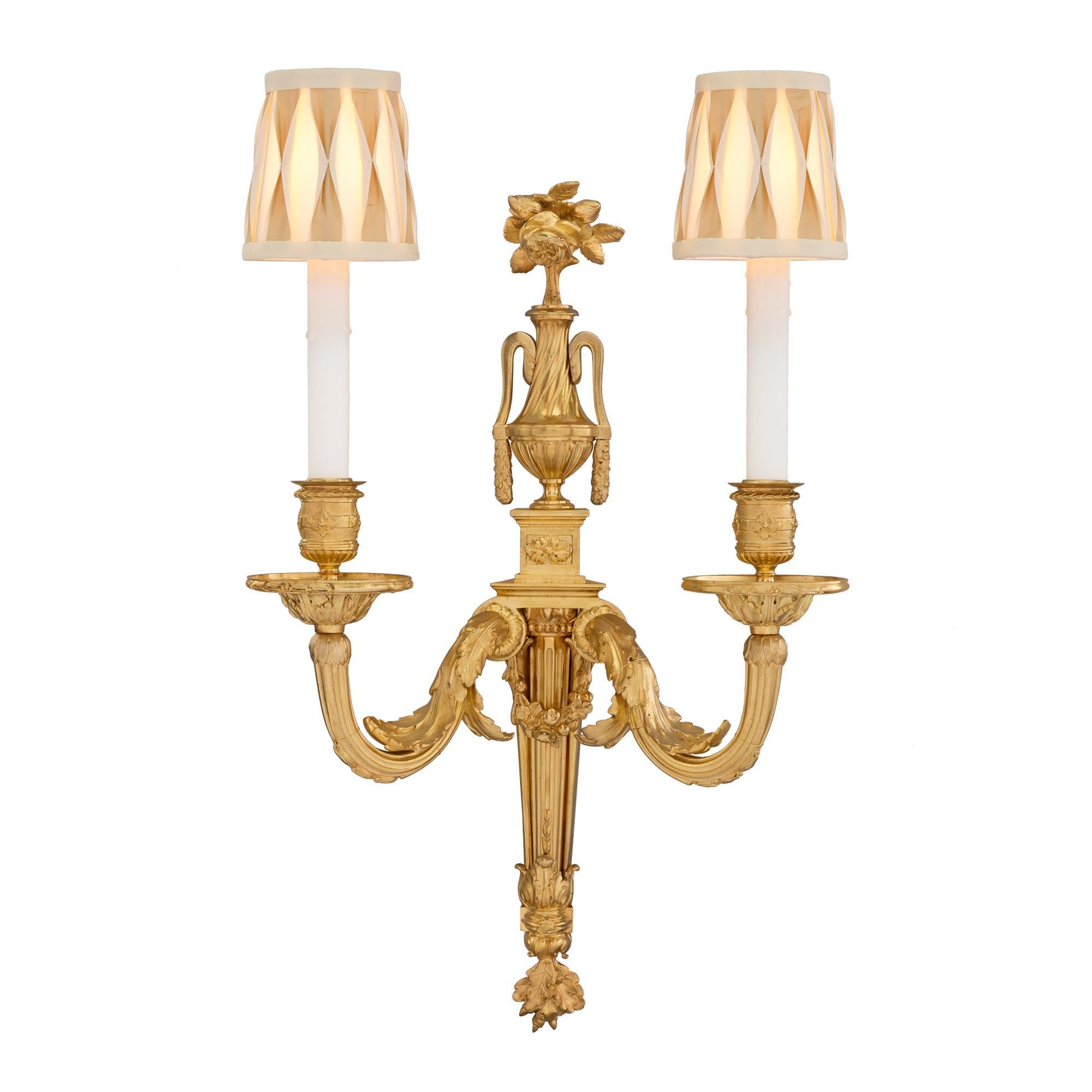 A handsome and statement making set of four French mid 19th century Louis XVI st. two arm ormolu sconces. The sconces top central urns of prosperity with blossoming flowers all on square supports with decorative rosettes. At the center of each