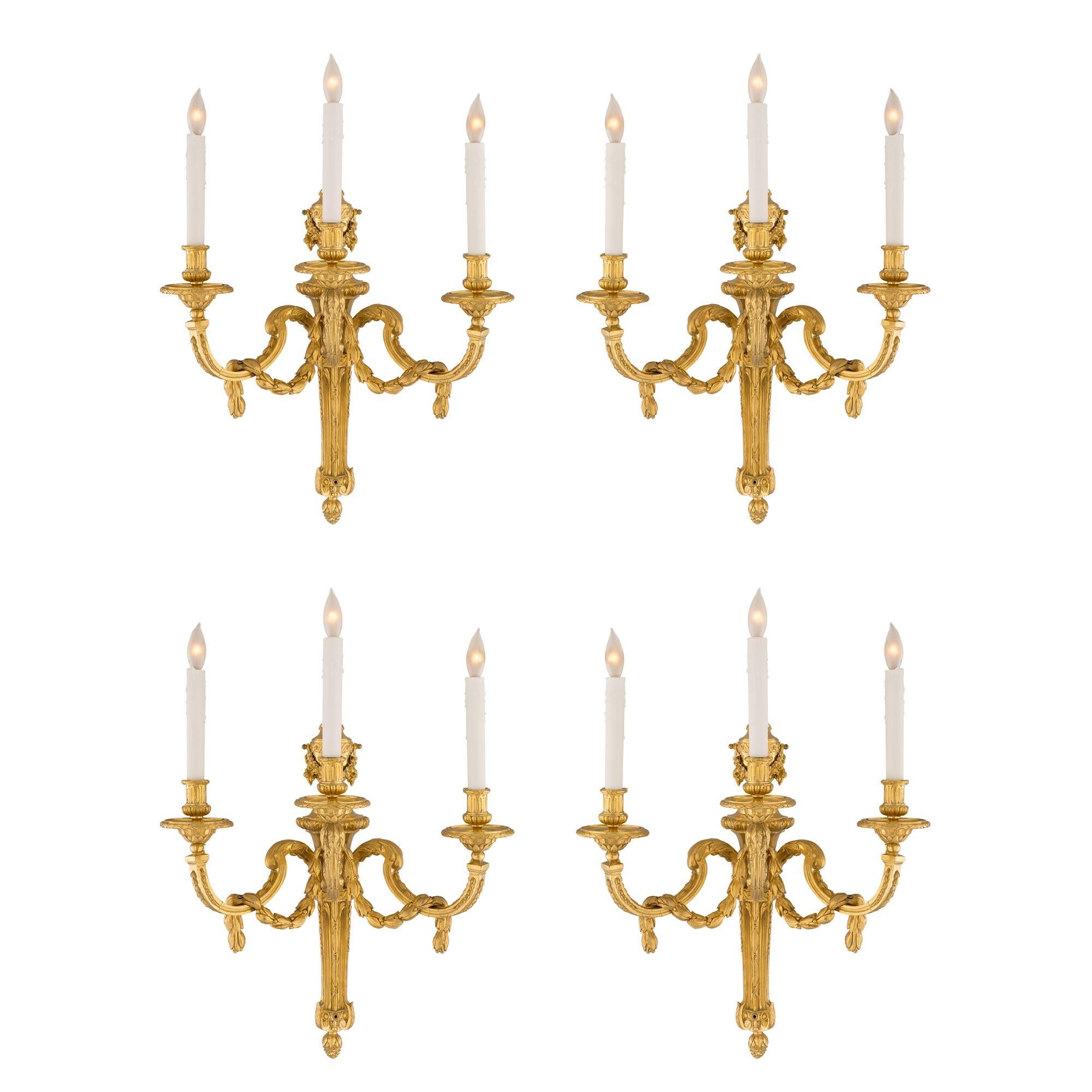 A sensational and grand scale set of four French mid-19th century Louis XVI style three light ormolu Bras de Lumière sconces. Each sconce is centered by a tapered, fluted and highly chased ormolu support with an inverted bottom acorn finial. The