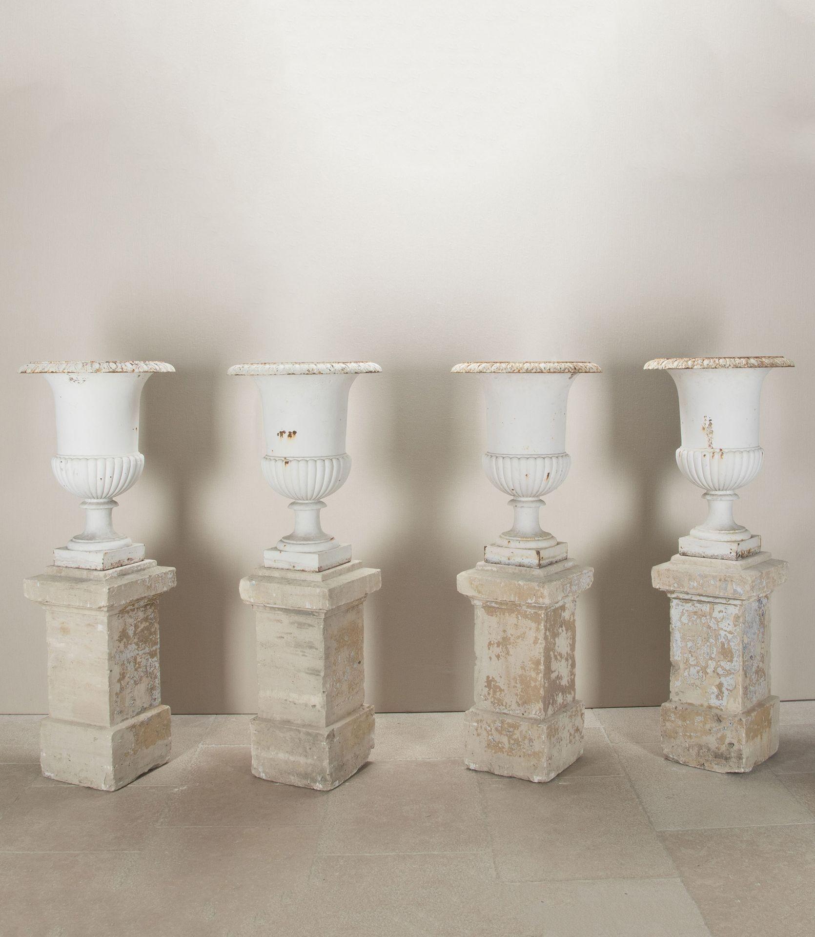 A finely cast set of four iron urns with egg and dart rims atop a plain body with a deeply gadrooned bowl in gently weathered white paint. The urns sit atop associated 19th Century carved stone plinths. One urn with chip to rim.