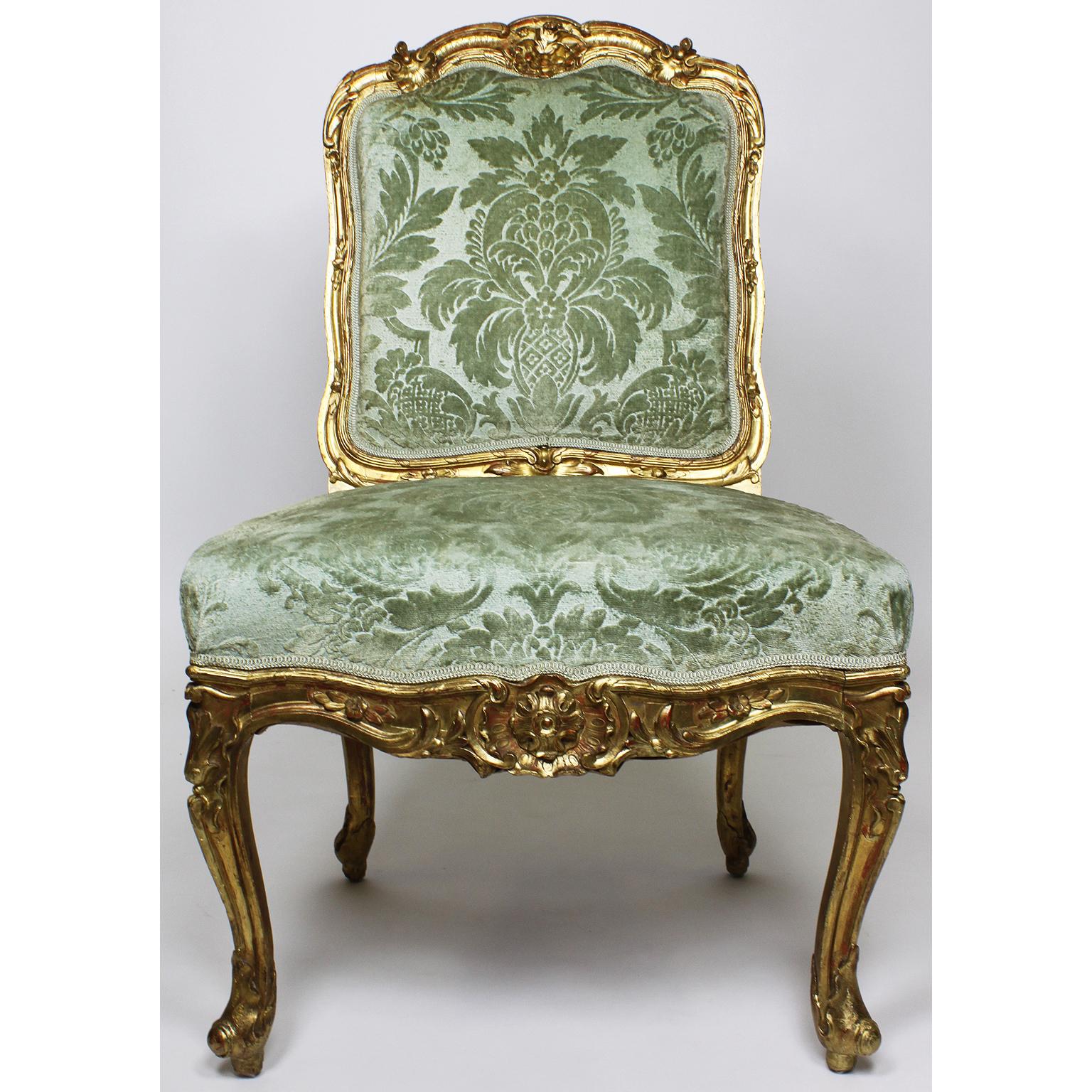 A fine set of four French 19th century Louis XV Rococo style giltwood carved chaises side-chairs, the arched upholstered panelled backs with foliate scroll cresting and similar borders, raised on flower heads decorated cabriolet legs ending in
