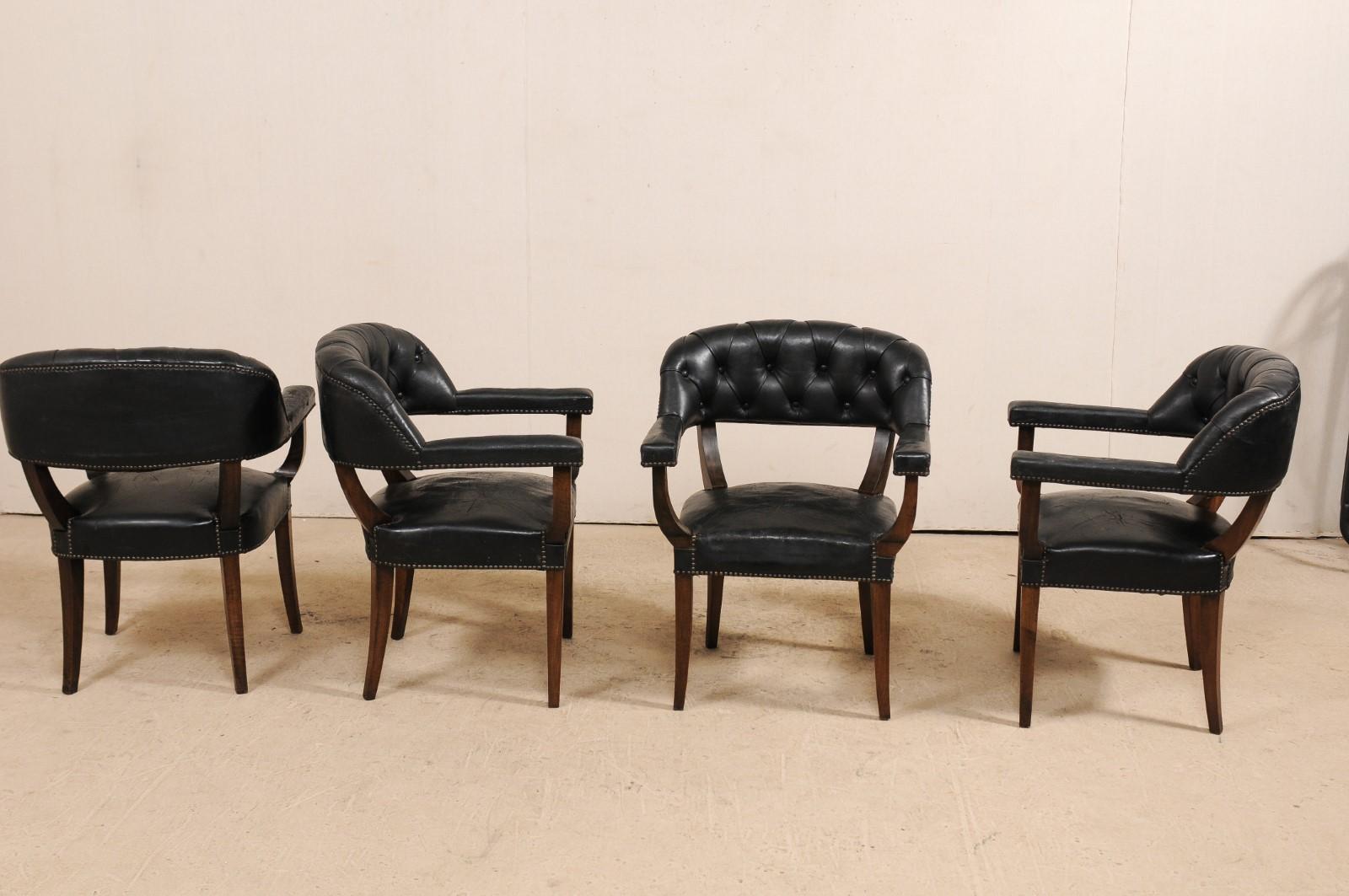 A handsome set of four French black leather arm chairs with rounded tub backs from the mid-20th century. This set of vintage chairs from France each feature their original tufted leather backs, straight arms and seat, presented upon wood saber legs.