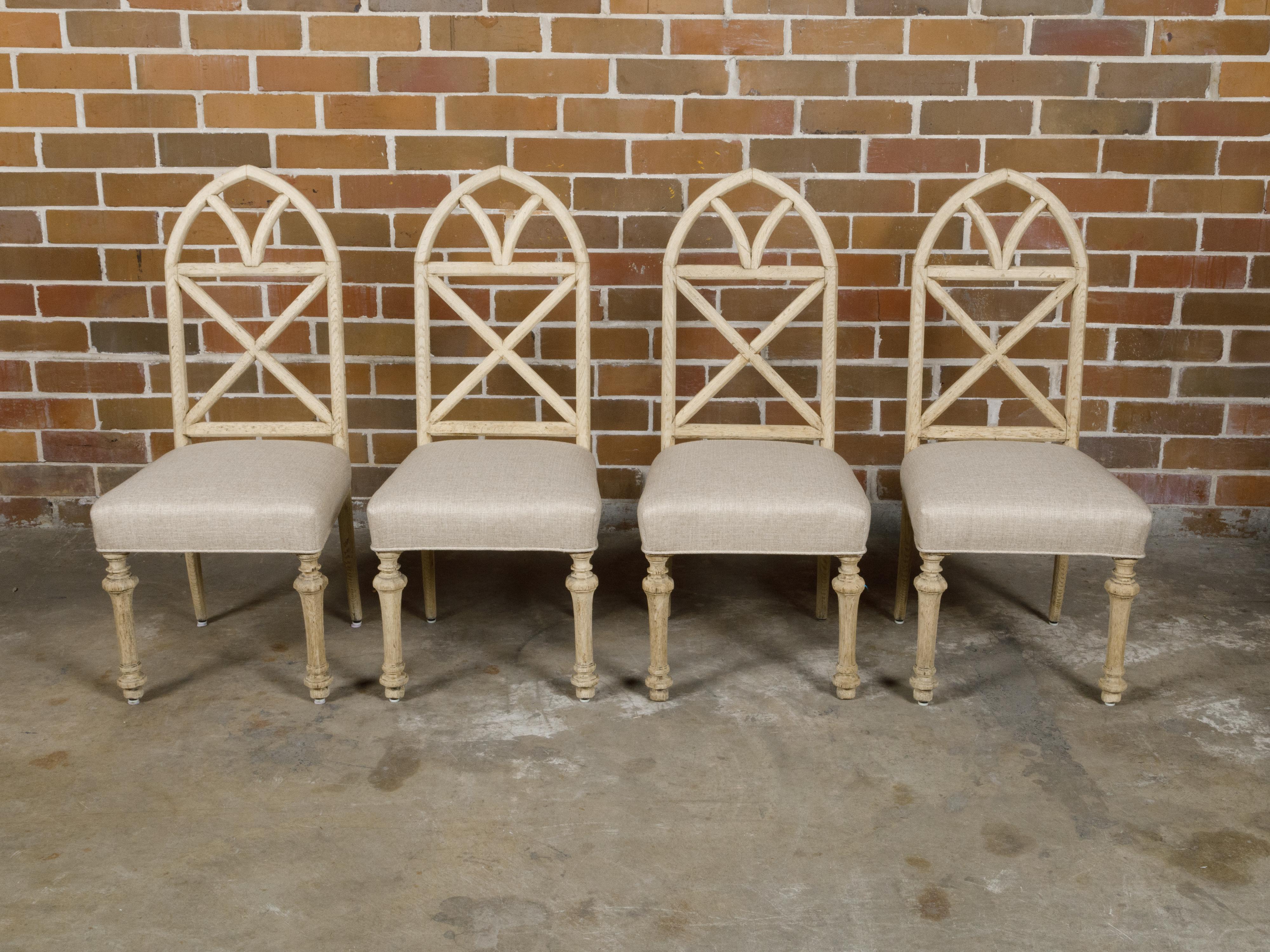 A set of four English Gothic Revival bleached oak dining room chairs with broken arch backs, faceted tapering legs and custom linen upholstery. This set of four English Gothic Revival dining room chairs, dating from the late 19th to early 20th