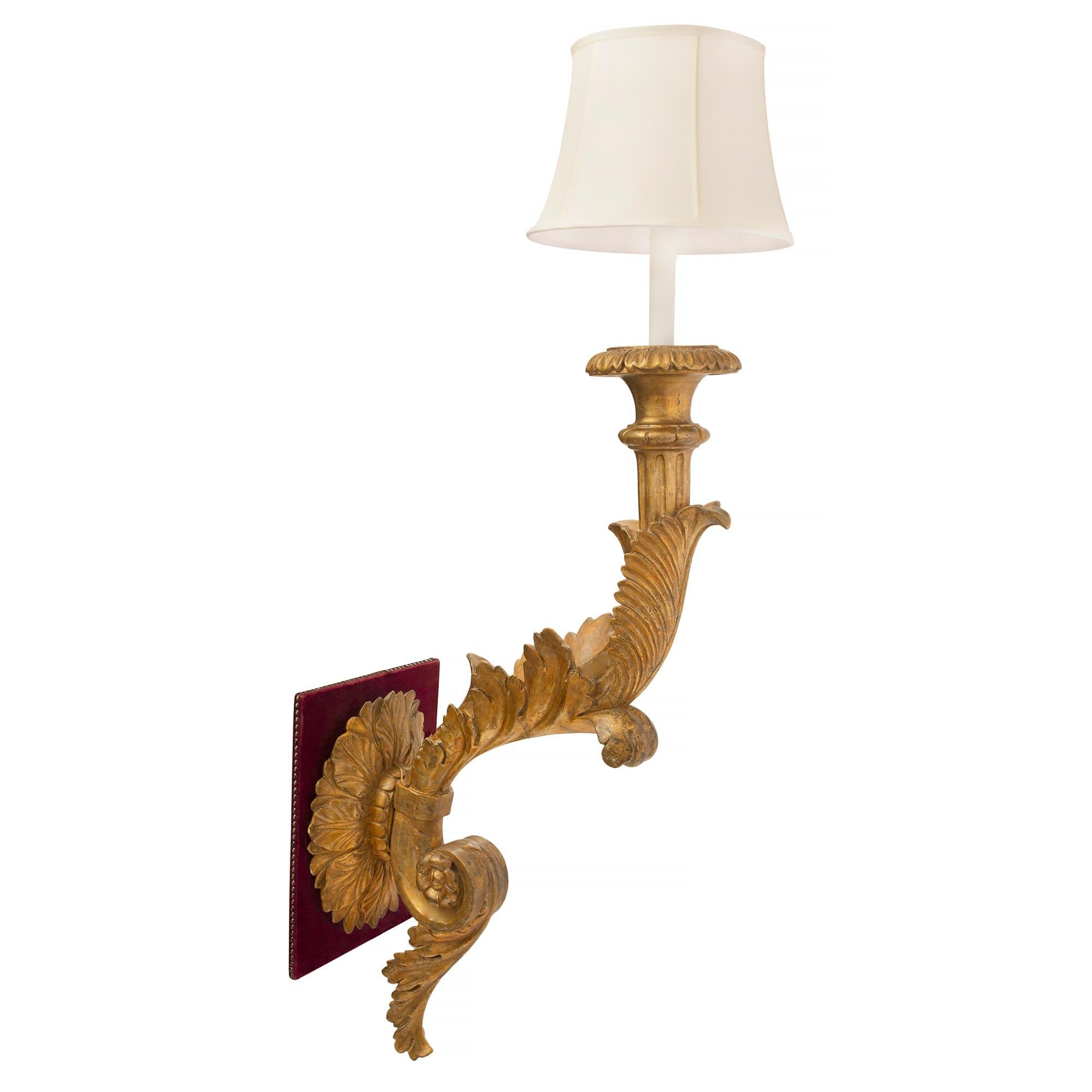 A handsome and large scale set of four Italian 18th century Louis XVI period giltwood electrified Bras de Lumière sconces. Each unique sconce is centered by an elegant and most decorative red velvet back plate finished with French nail heads and a