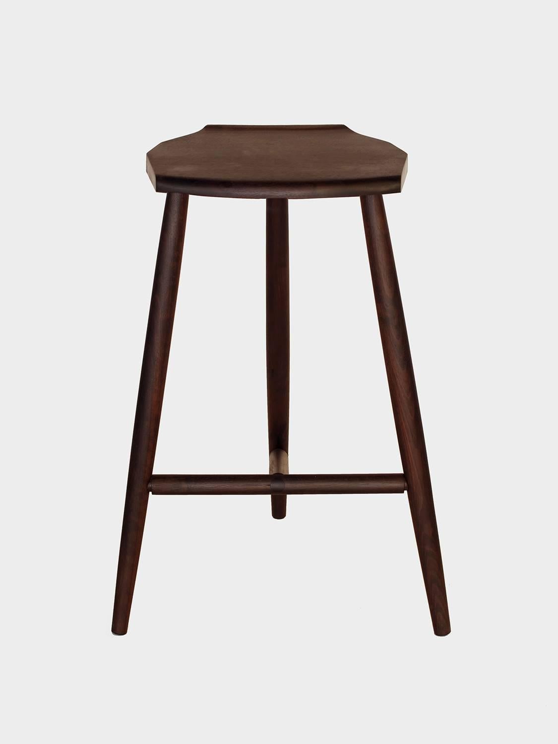 New York Heartwoods' ebonized walnut solid wood counter-height Jordan Stool is influenced by Shaker and Mid-Century design; created to be comfortable, lightweight, and easy to move; and features three turned legs, a unique hand-carved faceted seat,