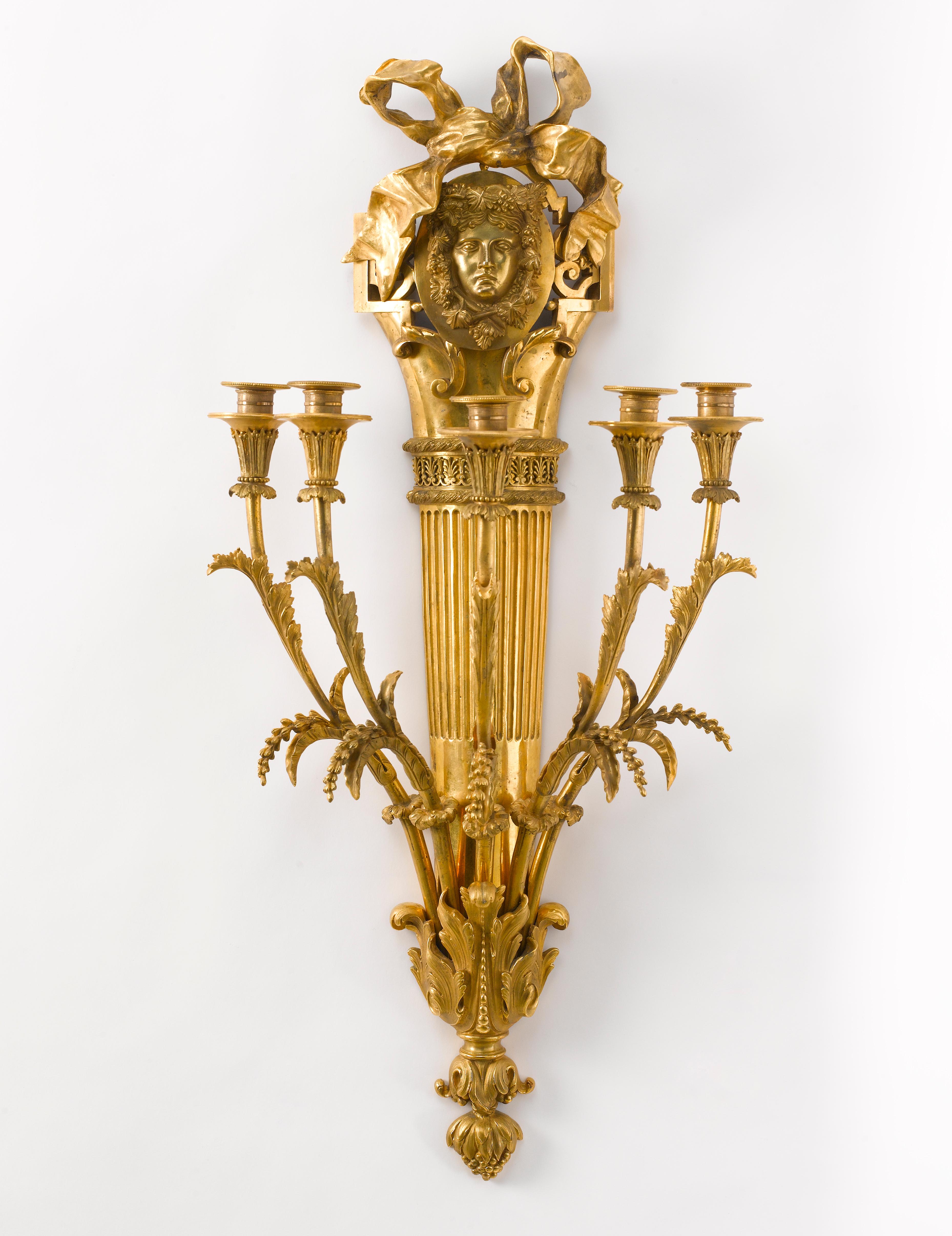 An impressive and rare set of four French or German gilt bronze five-branch scones. Each in the gout Grec (Greek style), with fluted backplate headed by a mask surrounded by wine leaves and surmounted by a large ribbon, issuing five