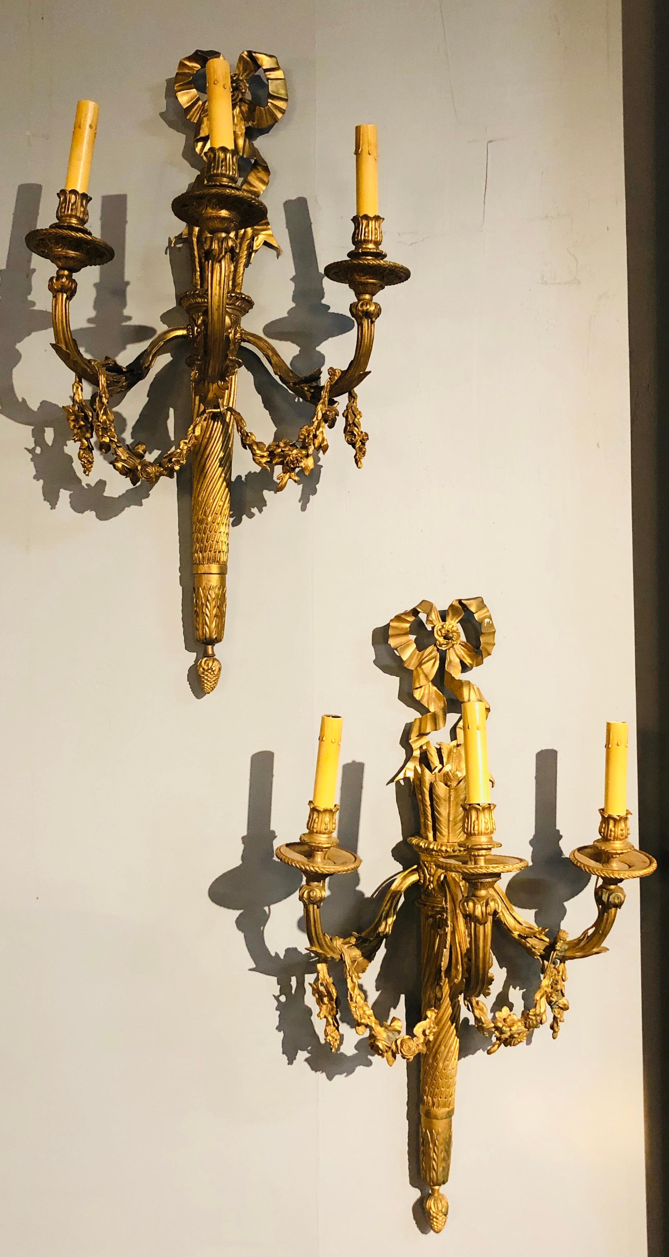 Set of four large and impressive Louis XVI style ornate three-light torch and ribbon form wall sconces. This listing is for two pair of these finely chiseled gilt bronze wall lights each newly wired taking three bulbs. The torch support having