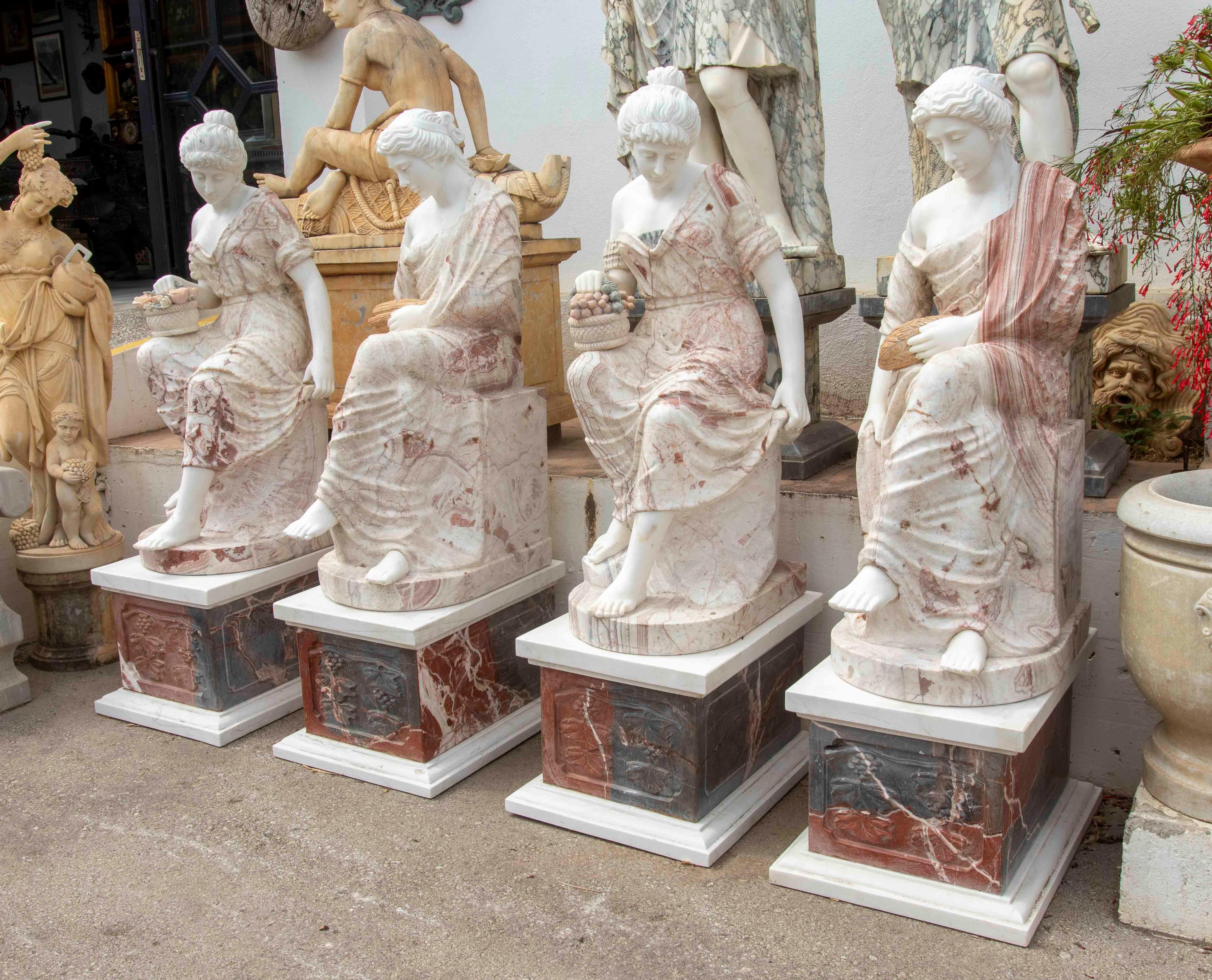 Impressive set of four carved marble sculptures depicting seated classical ladies, each holding different symbolic attributes such as baskets of flowers, fruit, and wheat ears. These pieces evoke the refinement of the Greco-Roman world, notable for