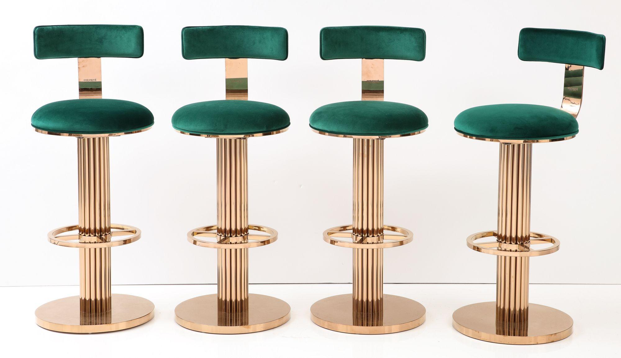A set of four barstools with a subtle rose gold finish and emerald velvet upholstery. This set is a reproduction of the Art Deco style Design For Leisure stools that were produced in the 1980s. Each base has a fluted design with a foot rest.
