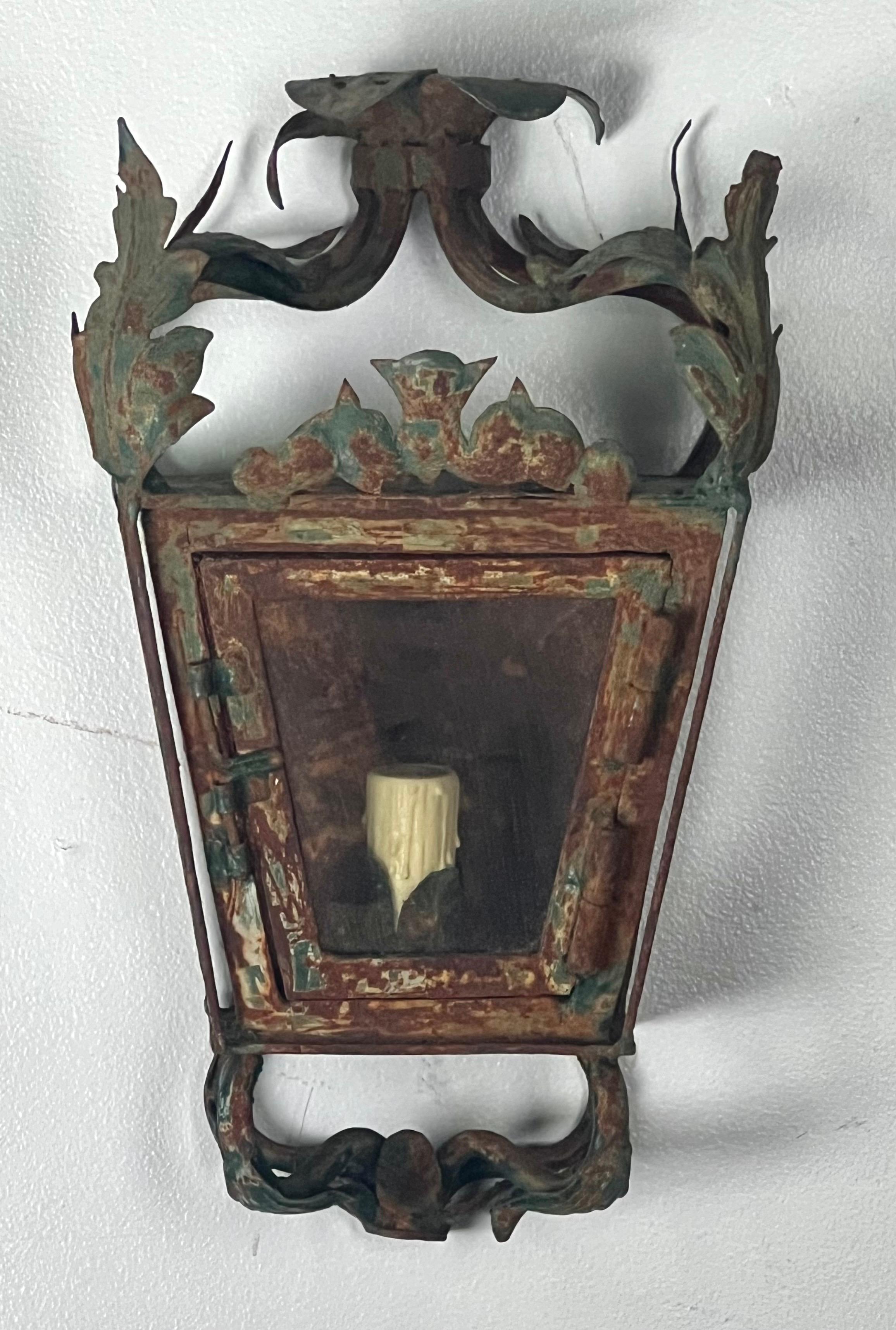 A set of four wrought iron vintage wall lanterns with a distinctly ornate design, featuring floral or foliate scrollwork.  They have been beautifully weathered,  The frames are made of wrought iron with a green patina and remnants of a rust finish. 