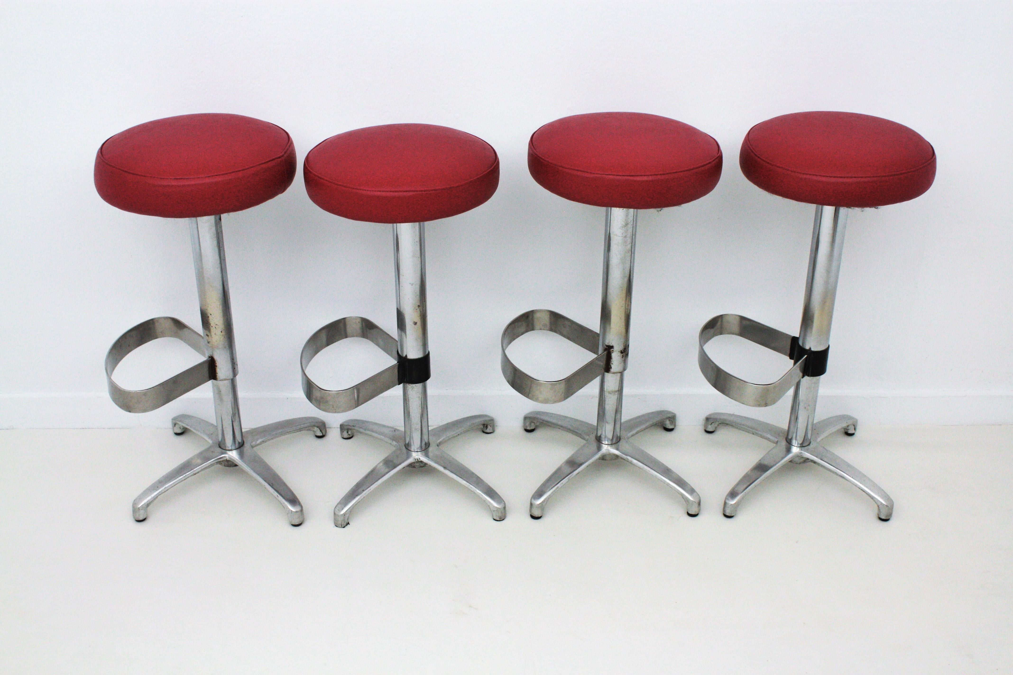 Set of four swivel bar stools in metal and red leatherette upholstered seats, Spain, 1970s.
These highly comfortable high barstools have a chromed steel structure with loop-shape footrest standing on a four-footed polished aluminium base. The soft