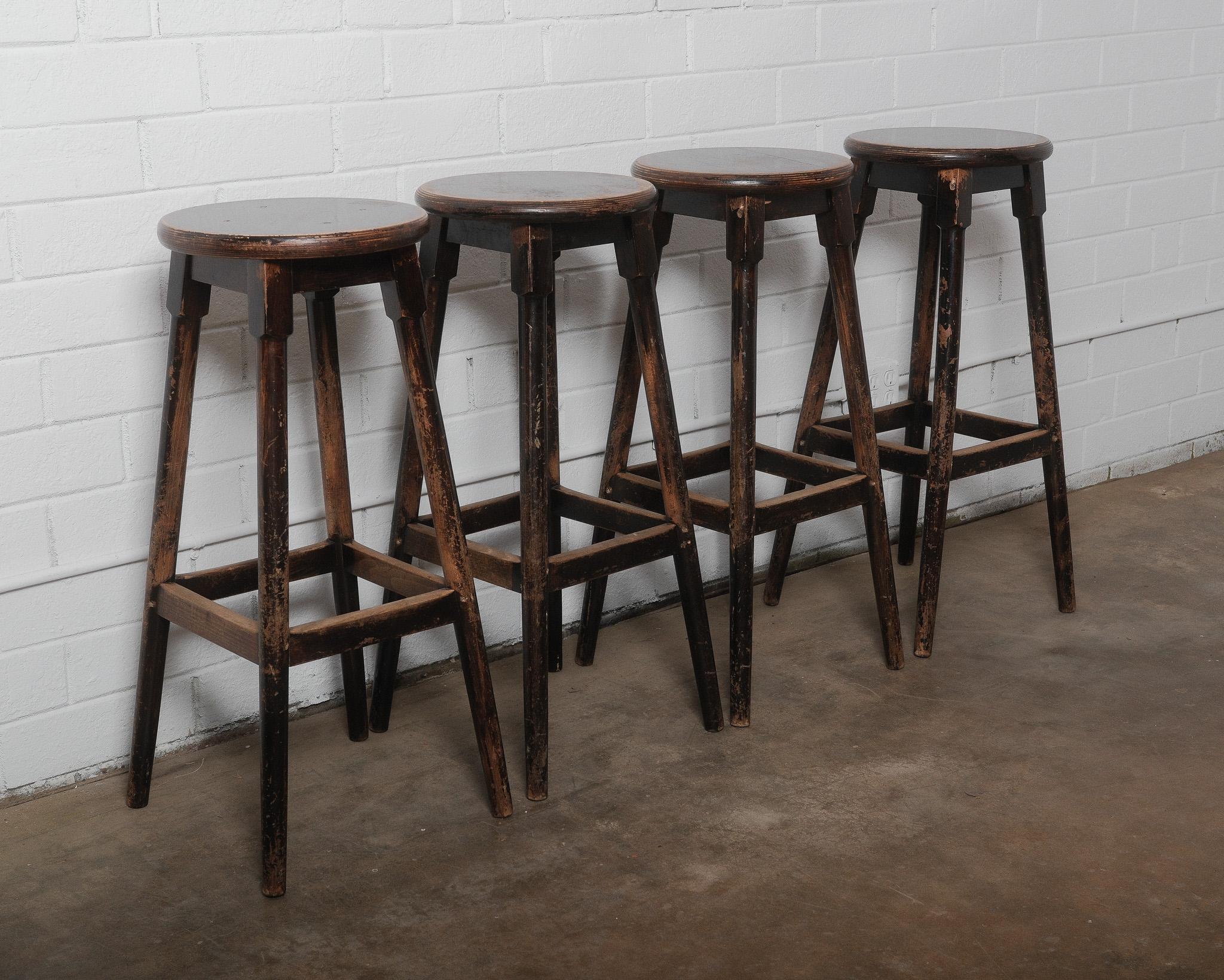 This set of four vintage wooden bar stools exudes rustic charm with their weathered dark finish and sturdy, classic design. Crafted from solid wood, these stools feature a round seat, splayed legs, and footrests for durability and comfort. The