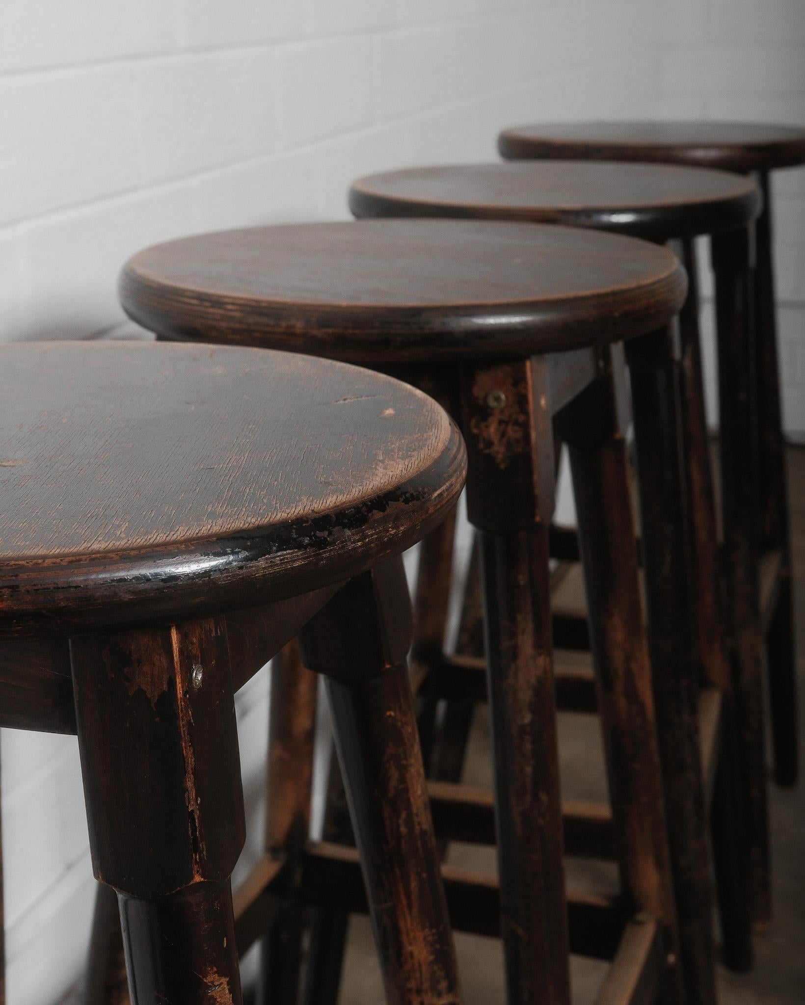 This set of four vintage wooden bar stools exudes rustic charm with their weathered dark finish and sturdy, classic design. Crafted from solid wood, these stools feature a round seat, splayed legs, and footrests for durability and comfort. The