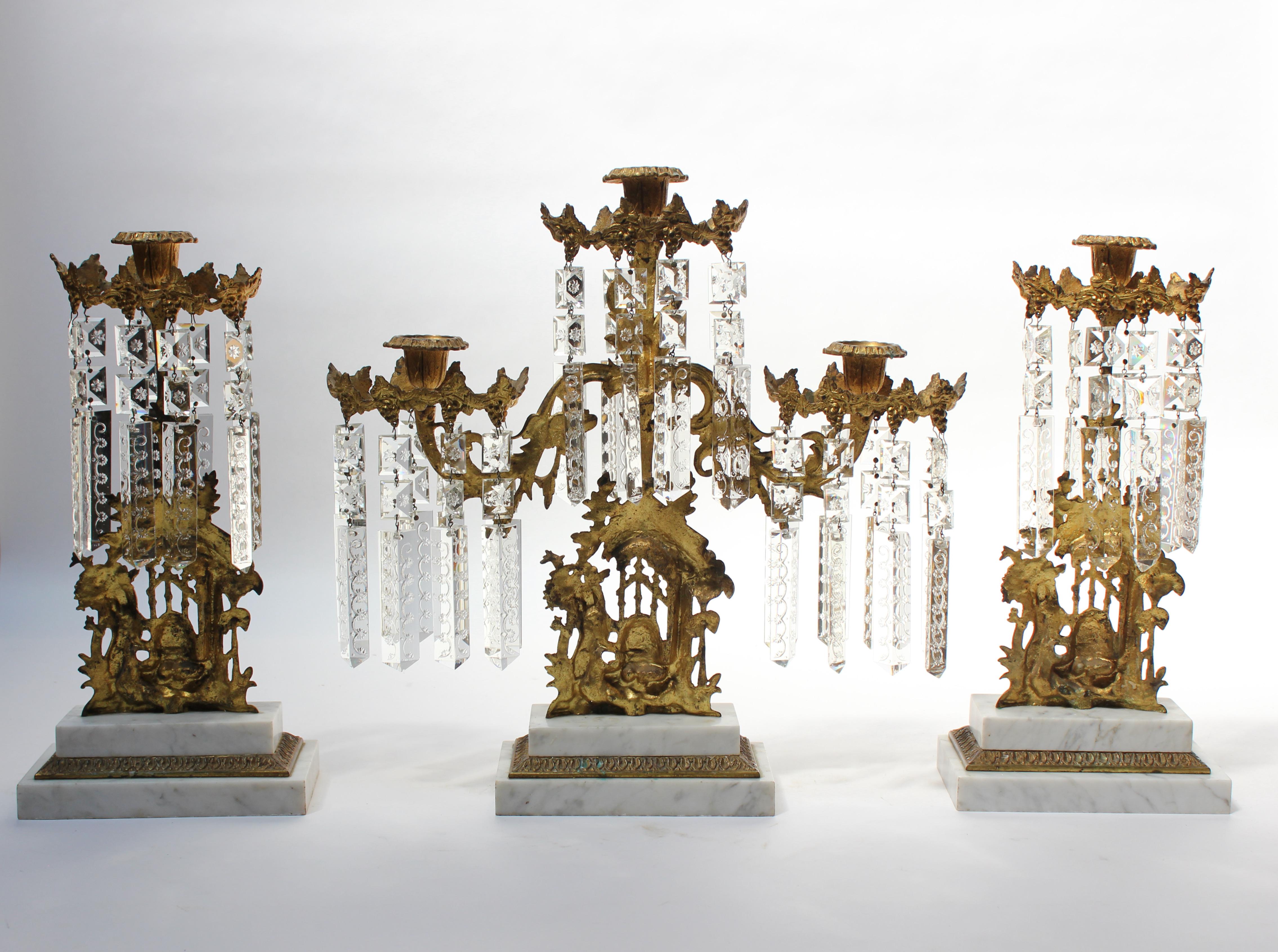 Rococo girandoles composed of gilt metal with stepped marble bases and etched lead-crystal prism decoration (ca. late 19th Century, France). The centerpiece candelabra features three branching arms accommodating three candles, while the smaller two