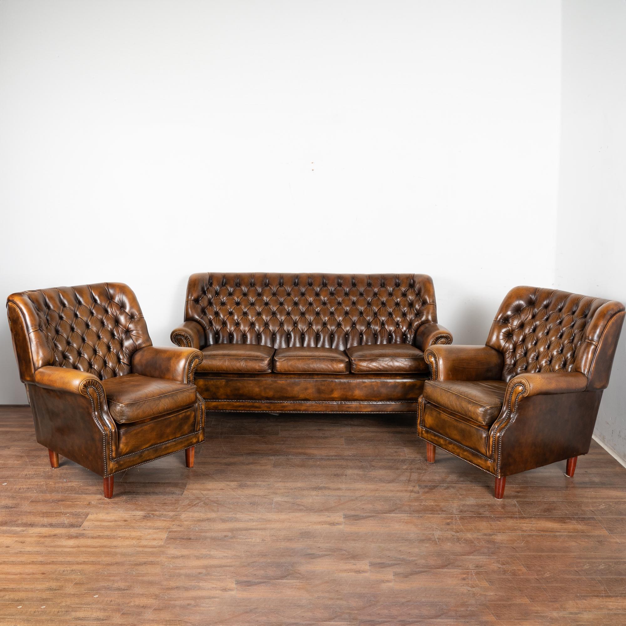 Set (3) vintage leather Chesterfield style three seat sofa and pair of club armchairs.
The mottled brown leather shows darker and lighter shades blending together is in good condition and accented by brass nail heads and traditional button tufted