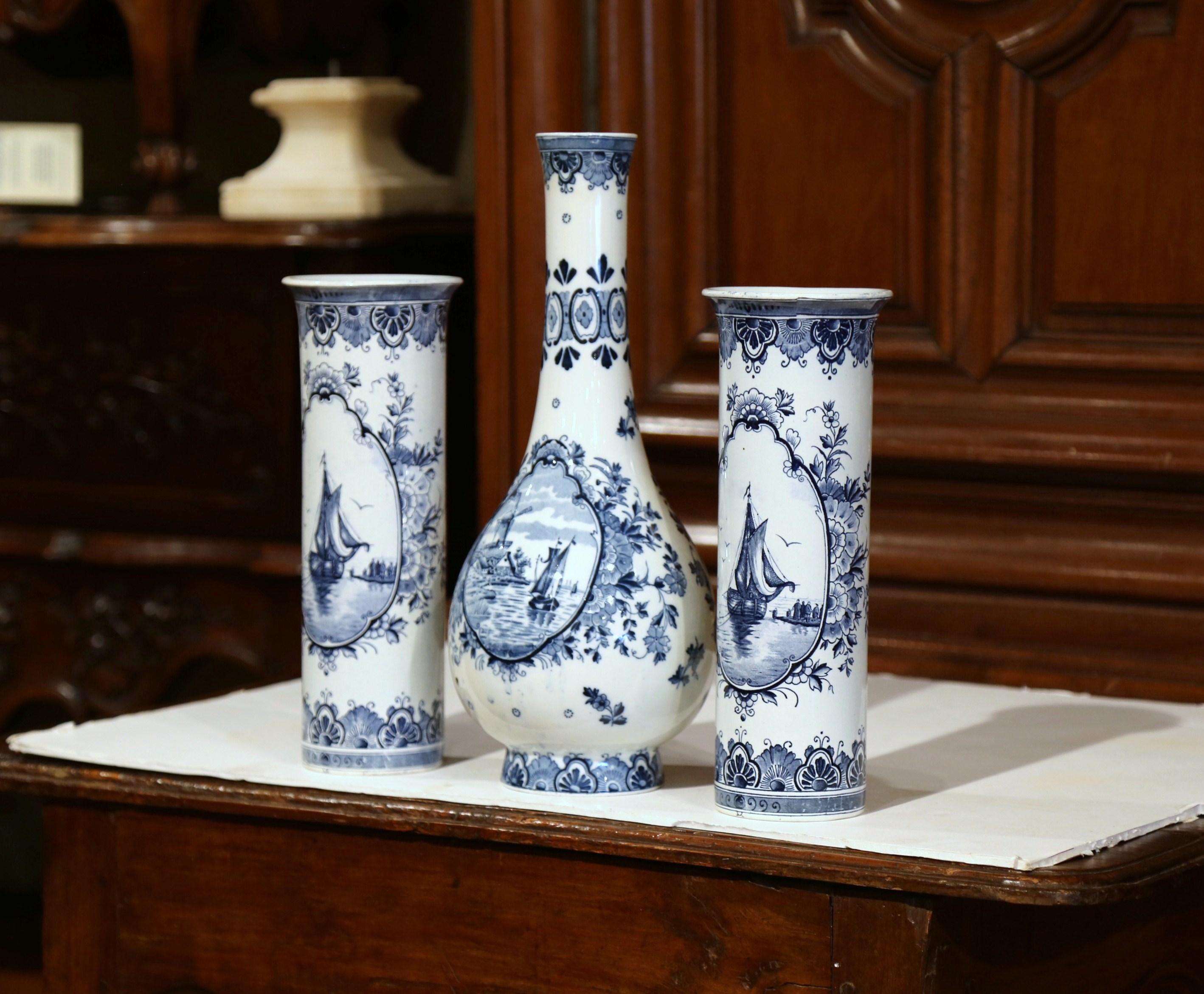 Decorate your mantel with this elegant set of porcelain Delft vases; crafted circa 1950, each hand-painted vase features a center sailboat on water medallion, embellished by floral decor in the blue and white palette. The tall center vase also