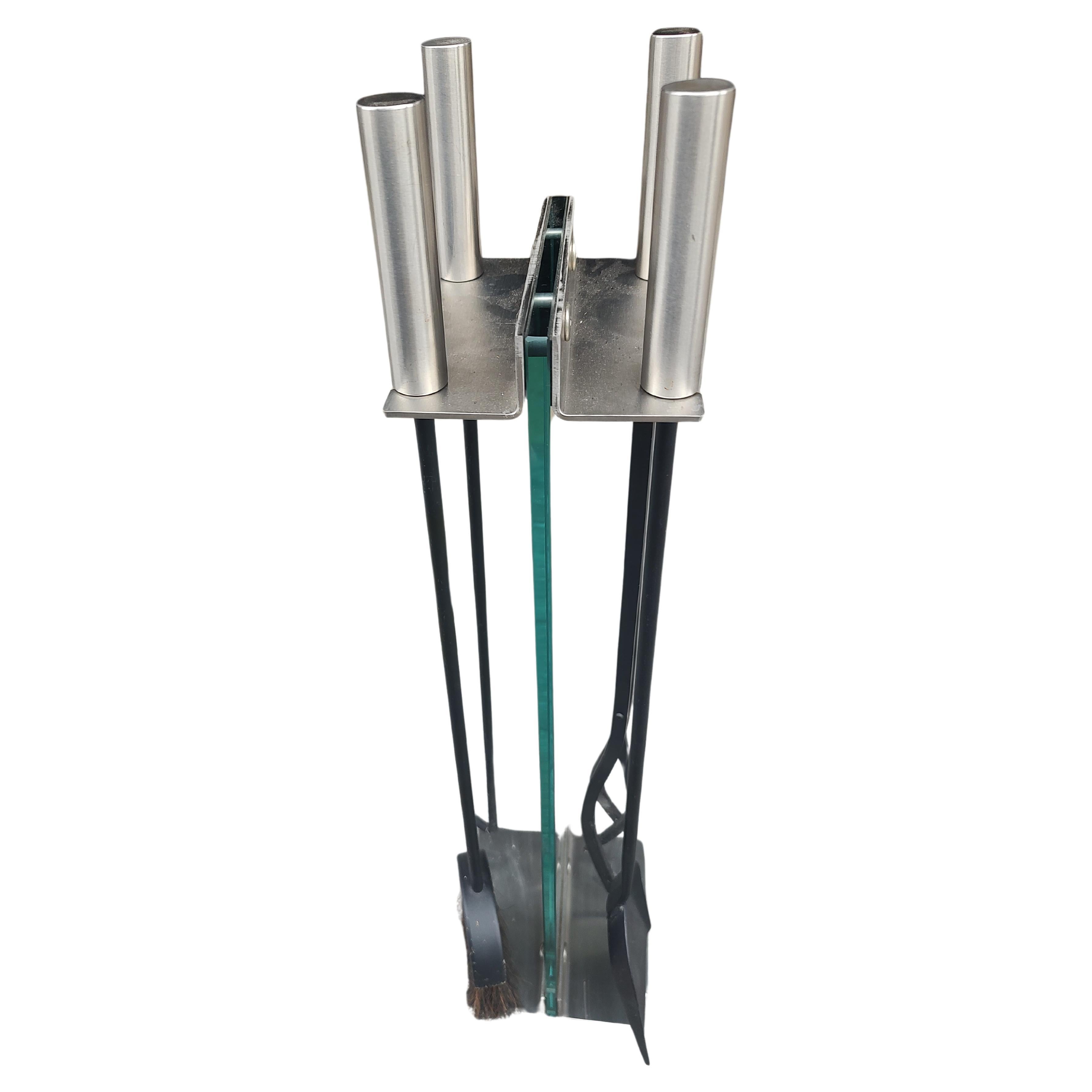 Fabuloso conjunto sencillo y elegante de herramientas para chimenea con mangos de acero inoxidable y un soporte cuya pieza central es un trozo de cristal. La parte superior e inferior del soporte también es de acero inoxidable. 5 piezas en total,