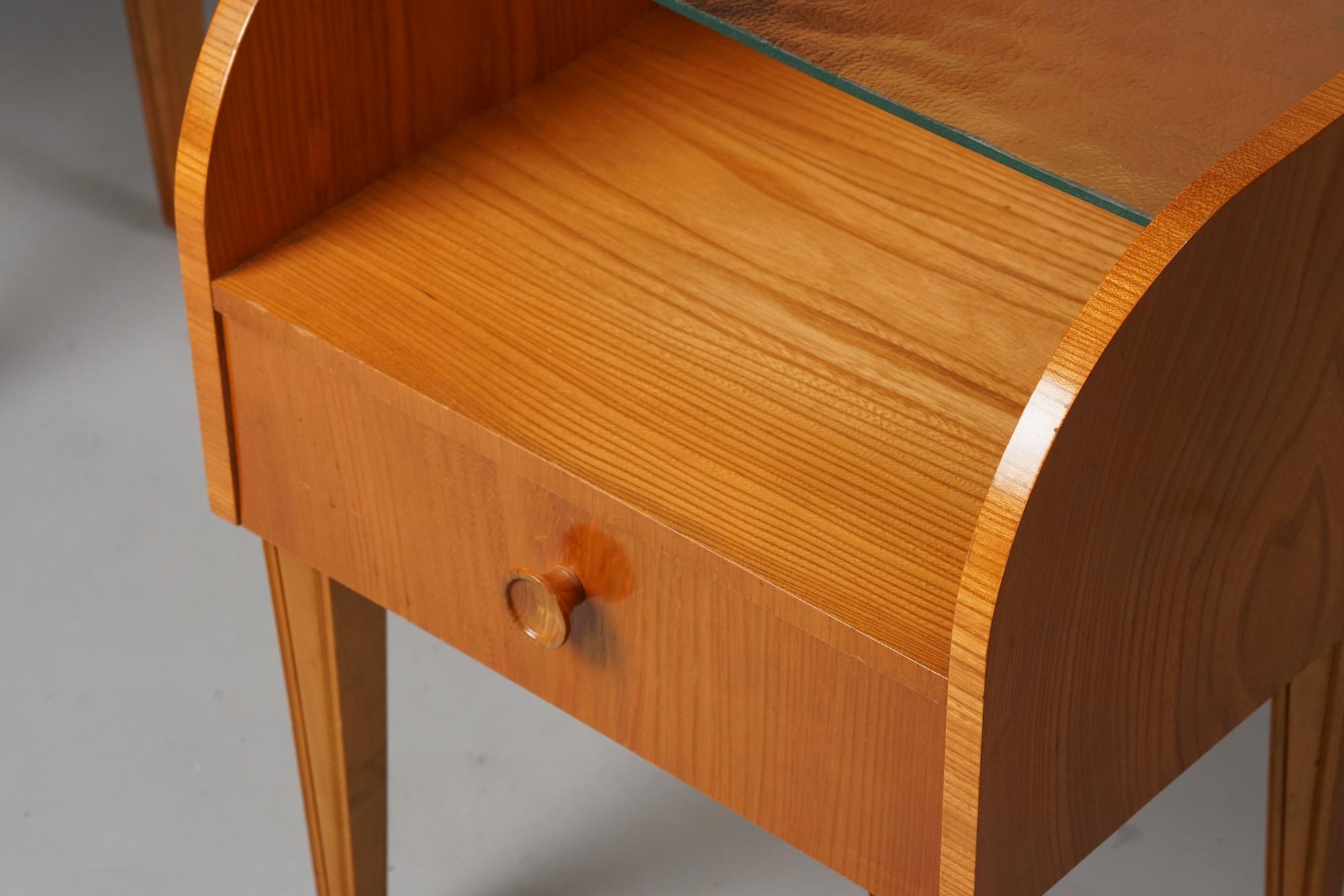 Set of night stands, designed by Margaret Nordman, manufactured by Keravan puusepäntehdas for Oy Stockmann Ab, 1930s. Elm frame with glass table top. Good vintage condition, patina consistent with age and use. The nightstand are sold as a set. 
