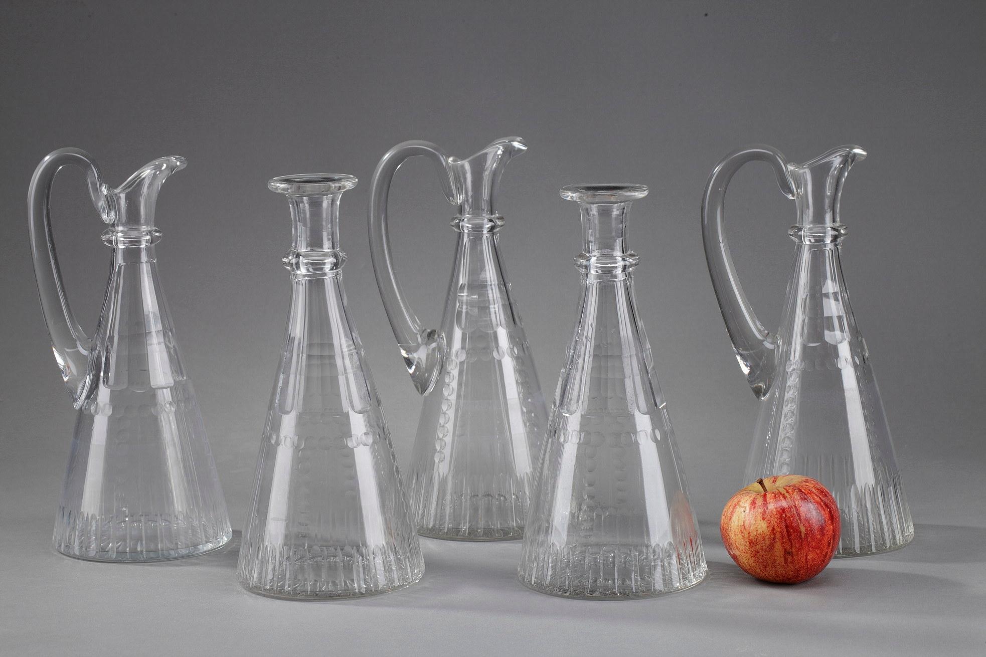 Set of three pitchers and two decanters in molded glass. The pitchers have a high handle that
connects the body and the lip of the containers. They end with a spout. The decanters have a thick
lip. All the glassware available has a unified