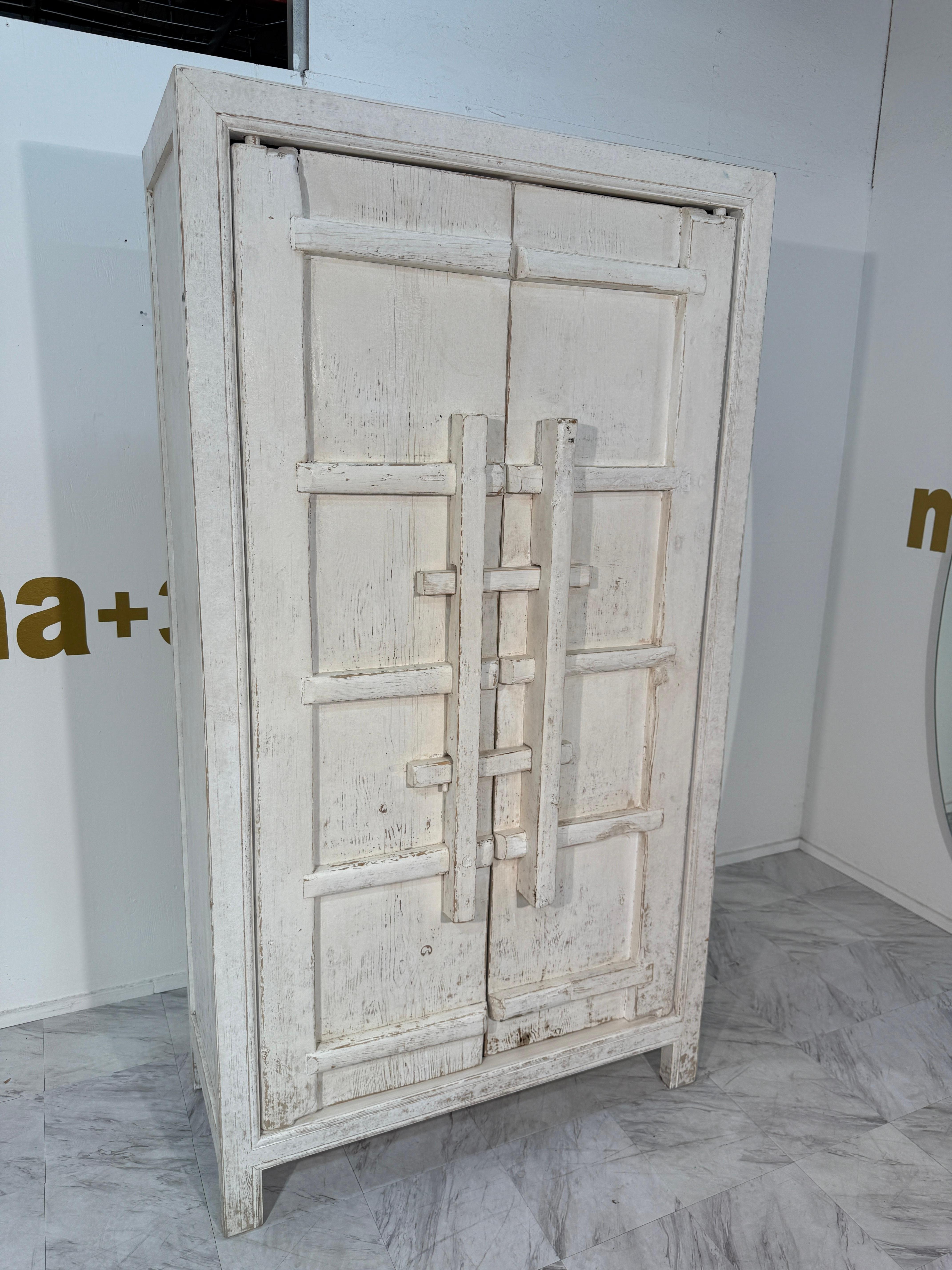 Incantevole armadio vintage in legno con una finitura bianca e distorta, che mostra audaci dettagli geometrici in rilievo sulle doppie porte. Il pezzo ha una forte presenza architettonica, con i suoi pannelli intagliati a griglia e le maniglie