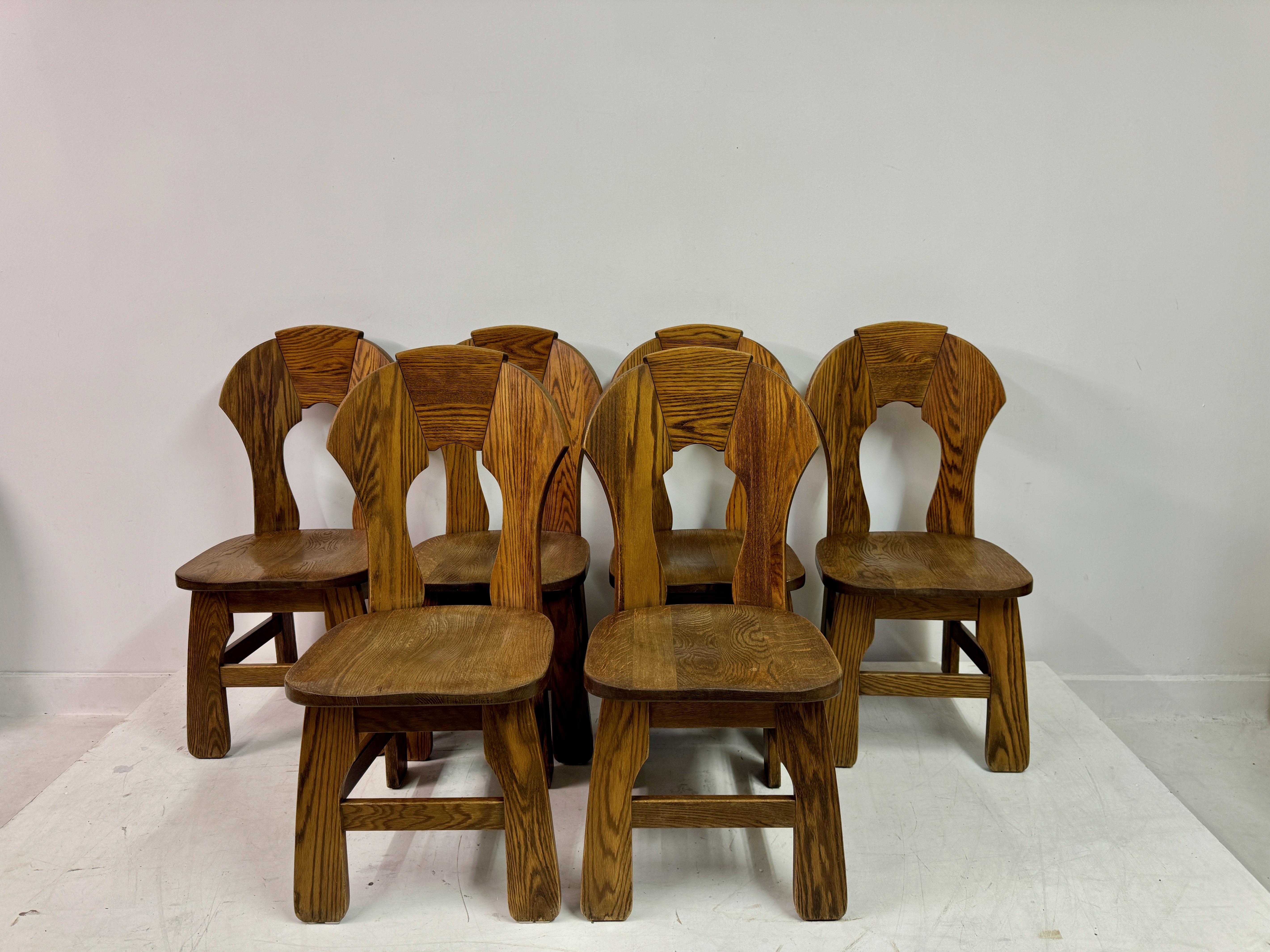 Questo set di sei sedie da pranzo belghe degli anni '70 esemplifica l'estetica audace e scultorea del design brutalista. Realizzata in rovere massiccio, ogni sedia ha una forma robusta e massiccia, con linee angolari e una presenza potente. Le sedie