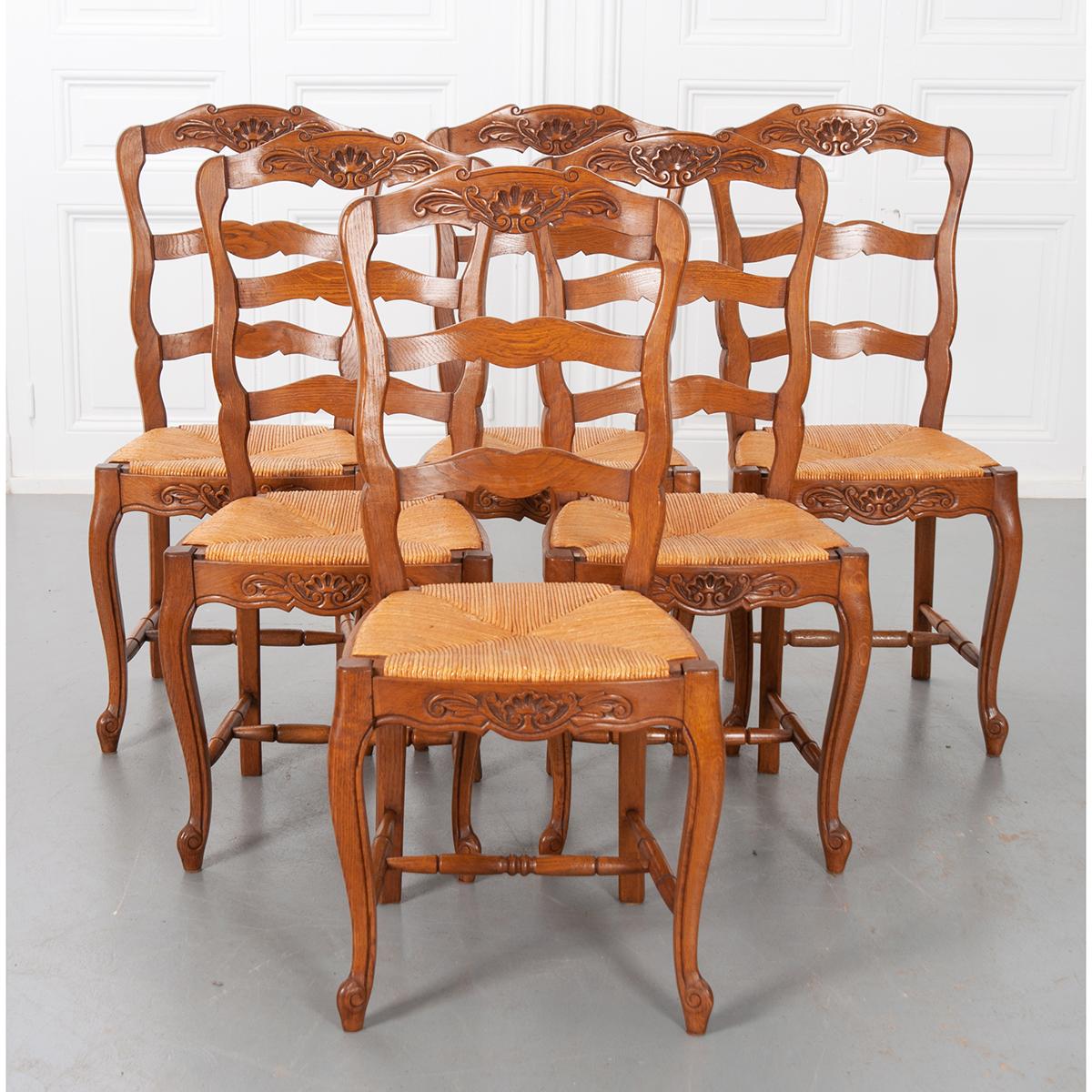 A set of six Provincial-style oak ladder back dining chairs with rush seats. The top and shaped front rails are each carved with beautiful shell cartouches. The chairs feature cabriole front legs that have scrolled escargot feet. The legs are braced