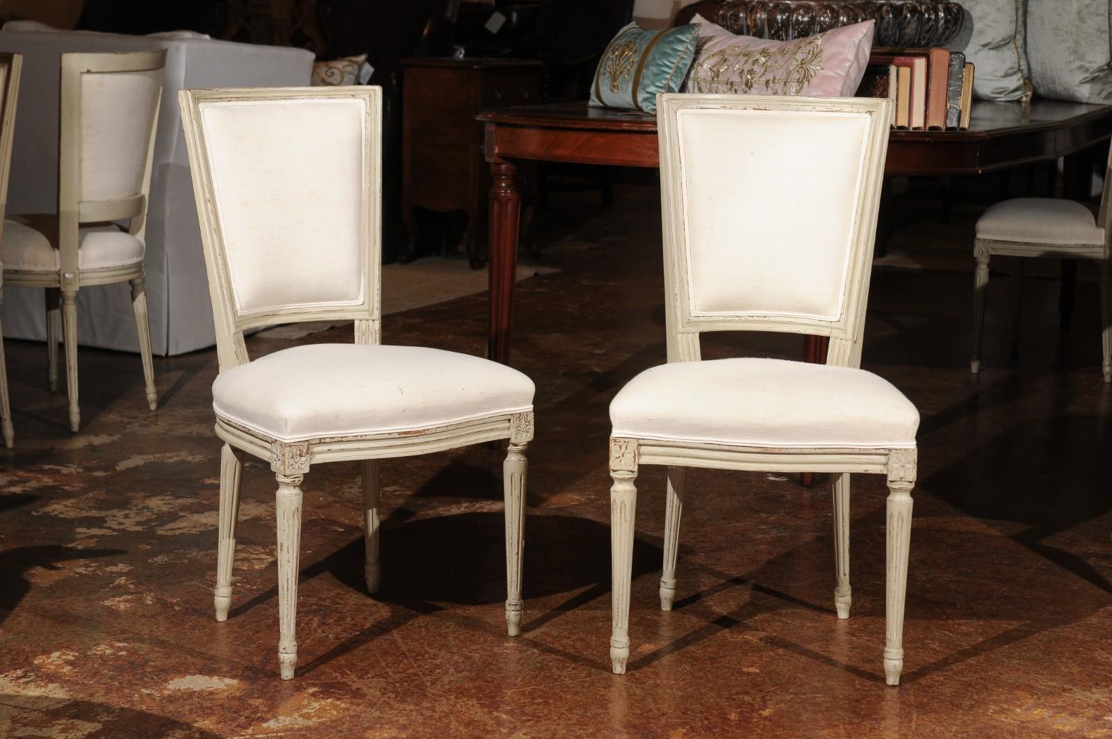 A set of six French Louis XVI style painted dining room side chairs from the early 20th century with fluted legs and new linen upholstery. Each of this set of French dining chairs features a trapezoidal curving back, resting on a double welt linen