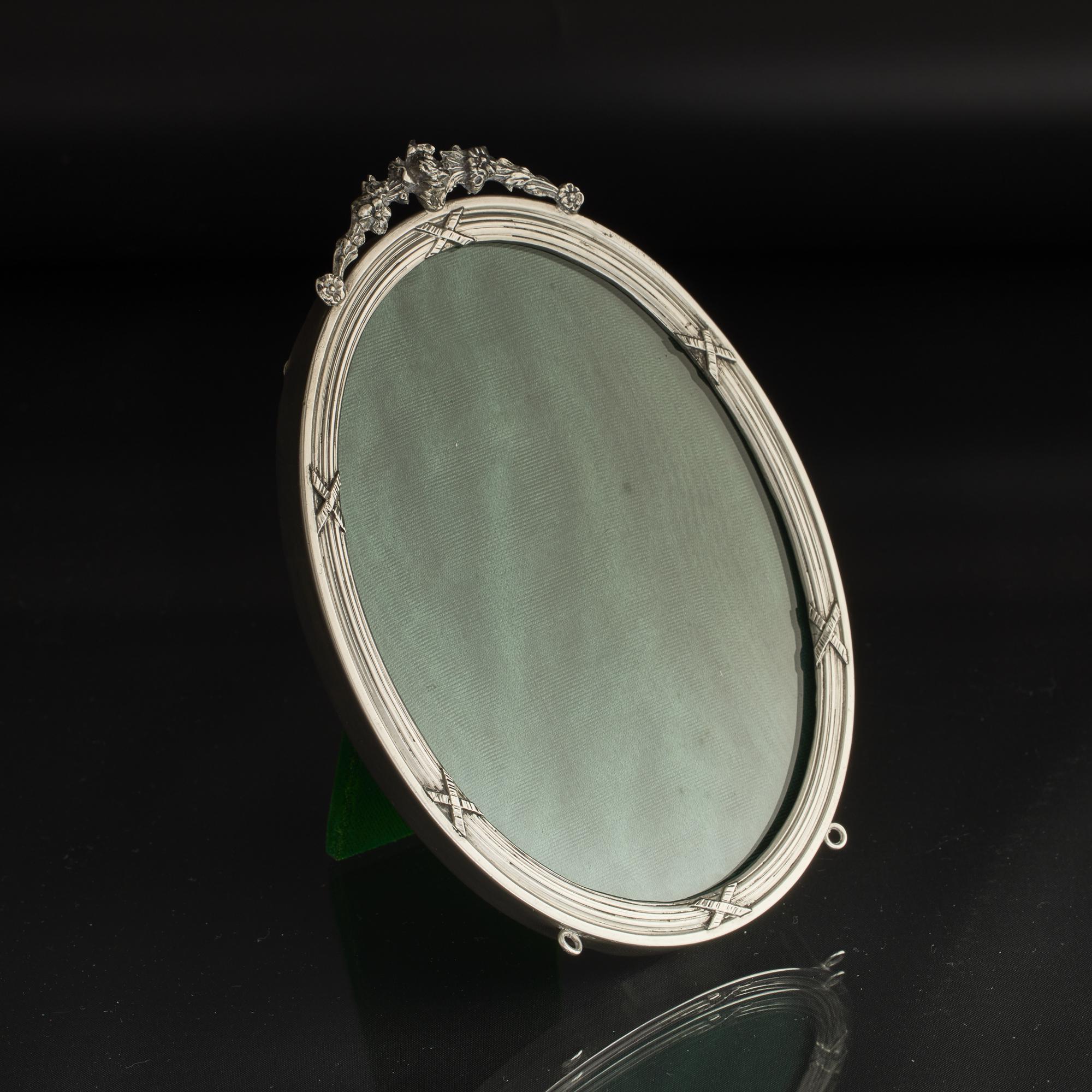 Marco de fotos antiguo ovalado de plata, de buena calidad, hecho a mano, con respaldo de terciopelo y caballete, de estilo eduardiano de principios del siglo XX. El marco plateado tiene un remate decorativo fundido con una rosa y otras flores,